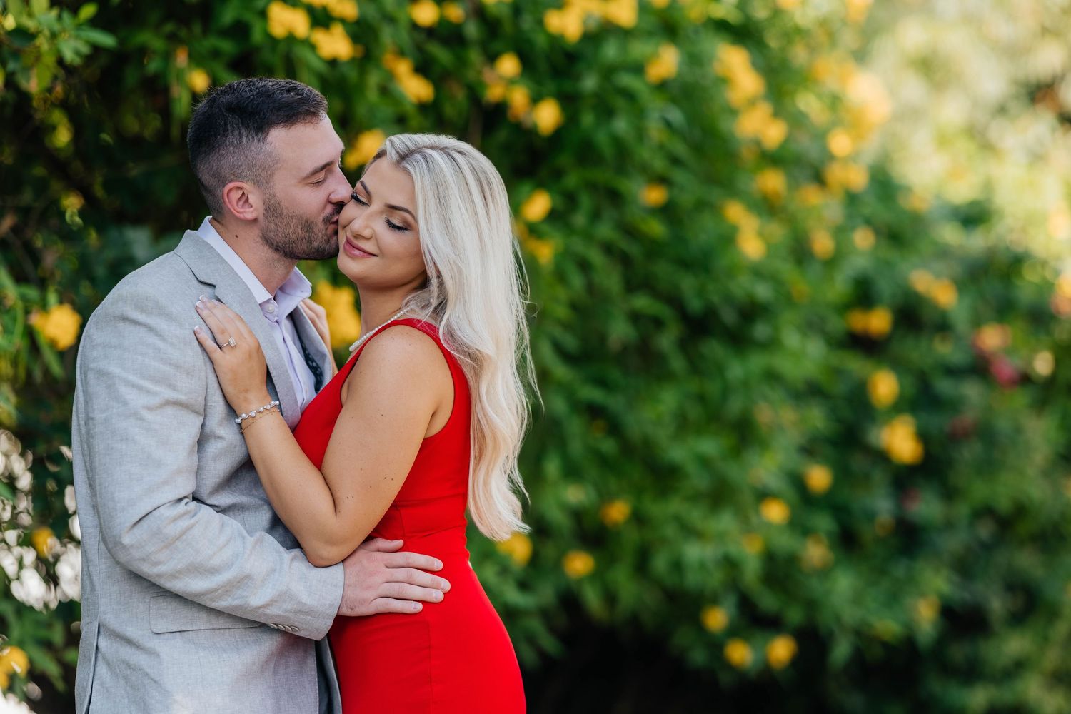 A romantic couple embraces outdoors against a backdrop of yellow flowers and greenery.