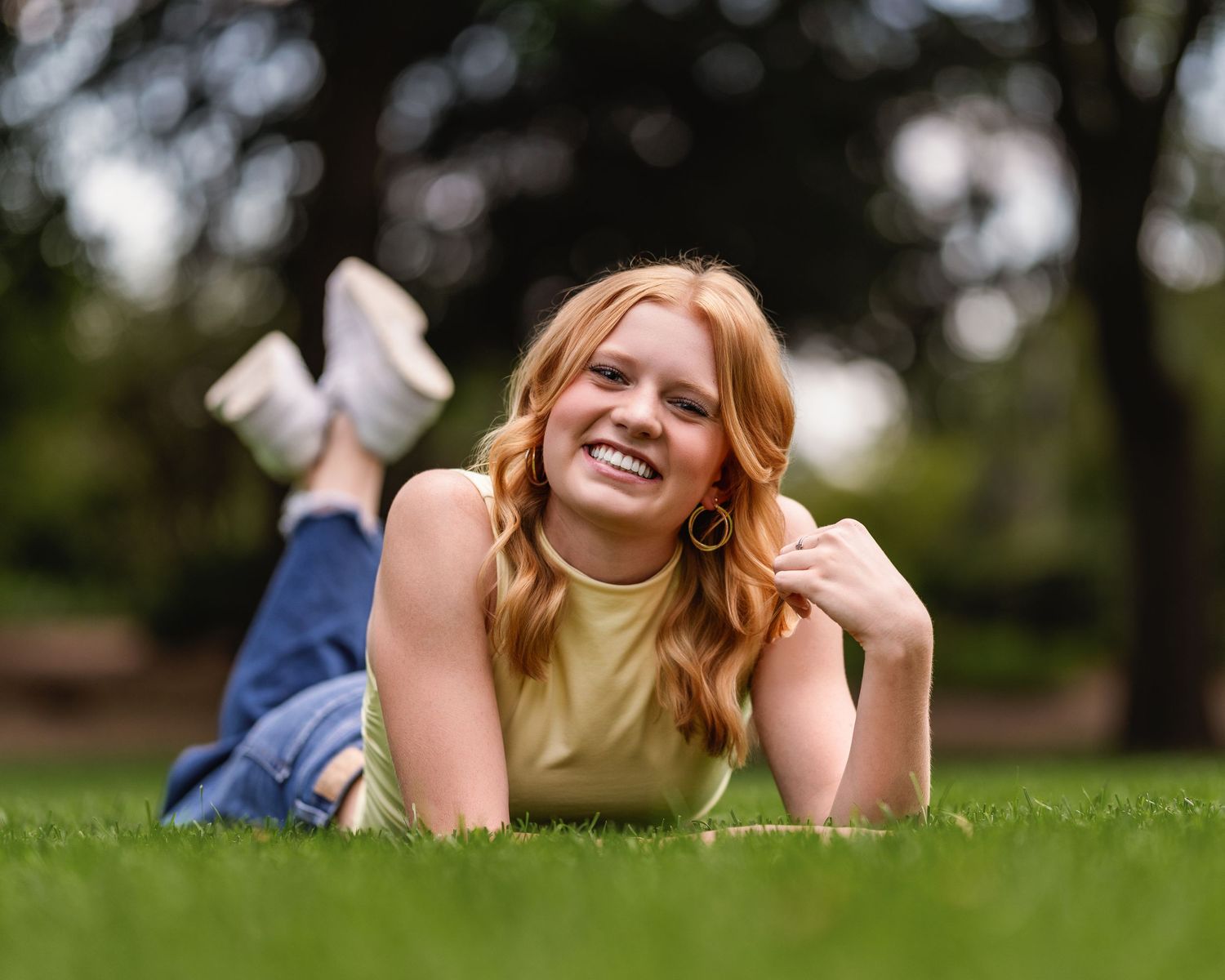 Smiling redhead teen girl lying on green grass in a park, wearing a yellow top and jeans, senior photo.