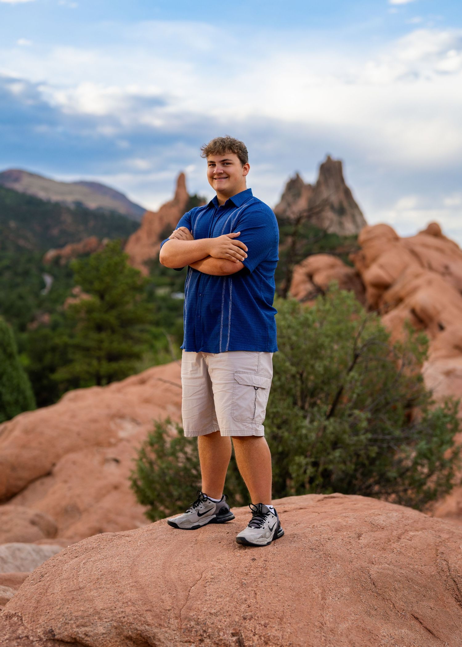 Young man in blue polo and khaki shorts stands arms crossed on red rocks at Garden of the Gods, Colorado.