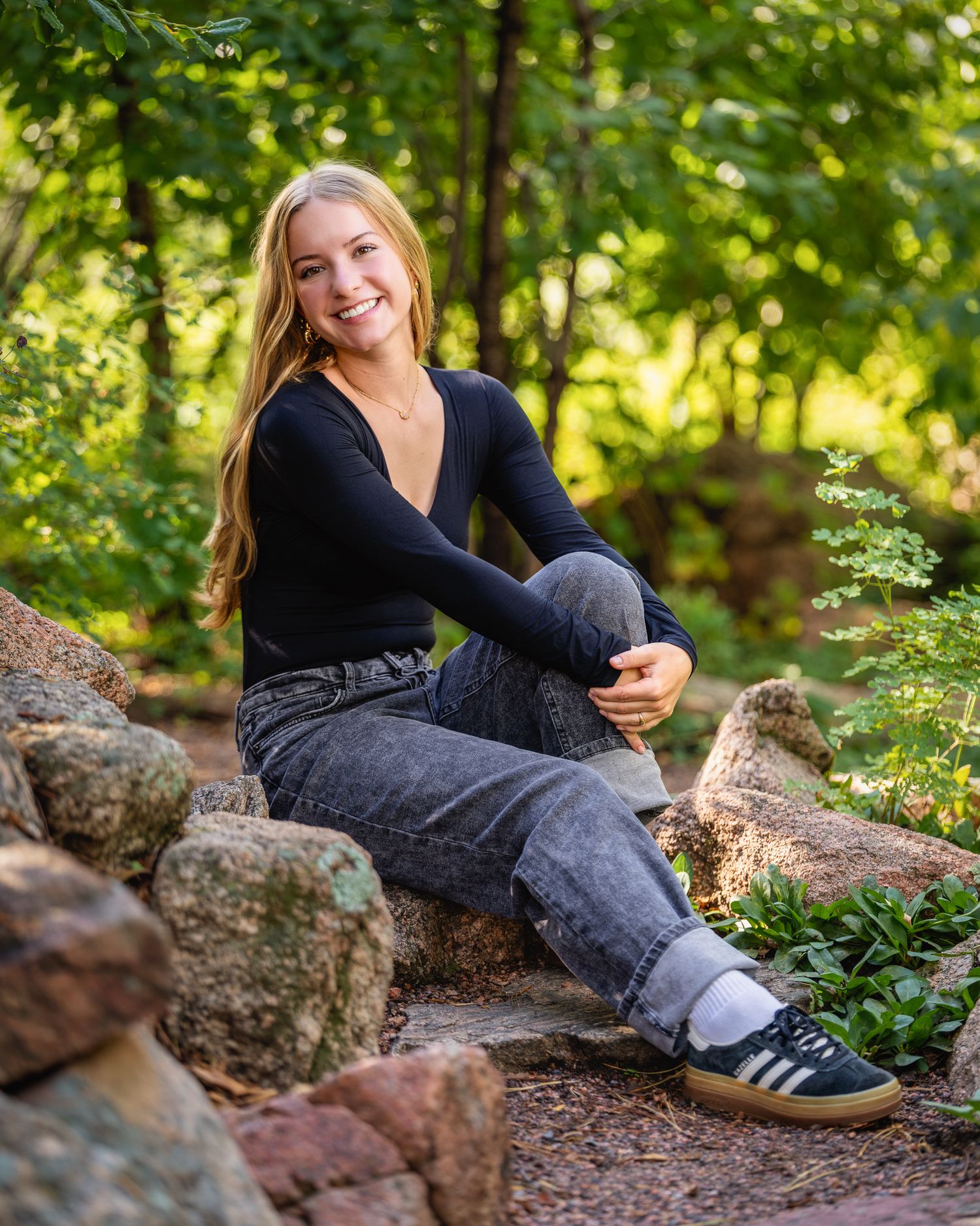 Young blonde woman smiling, sitting on rocks in a garden, wearing black top, gray jeans, and Adidas sneakers.
