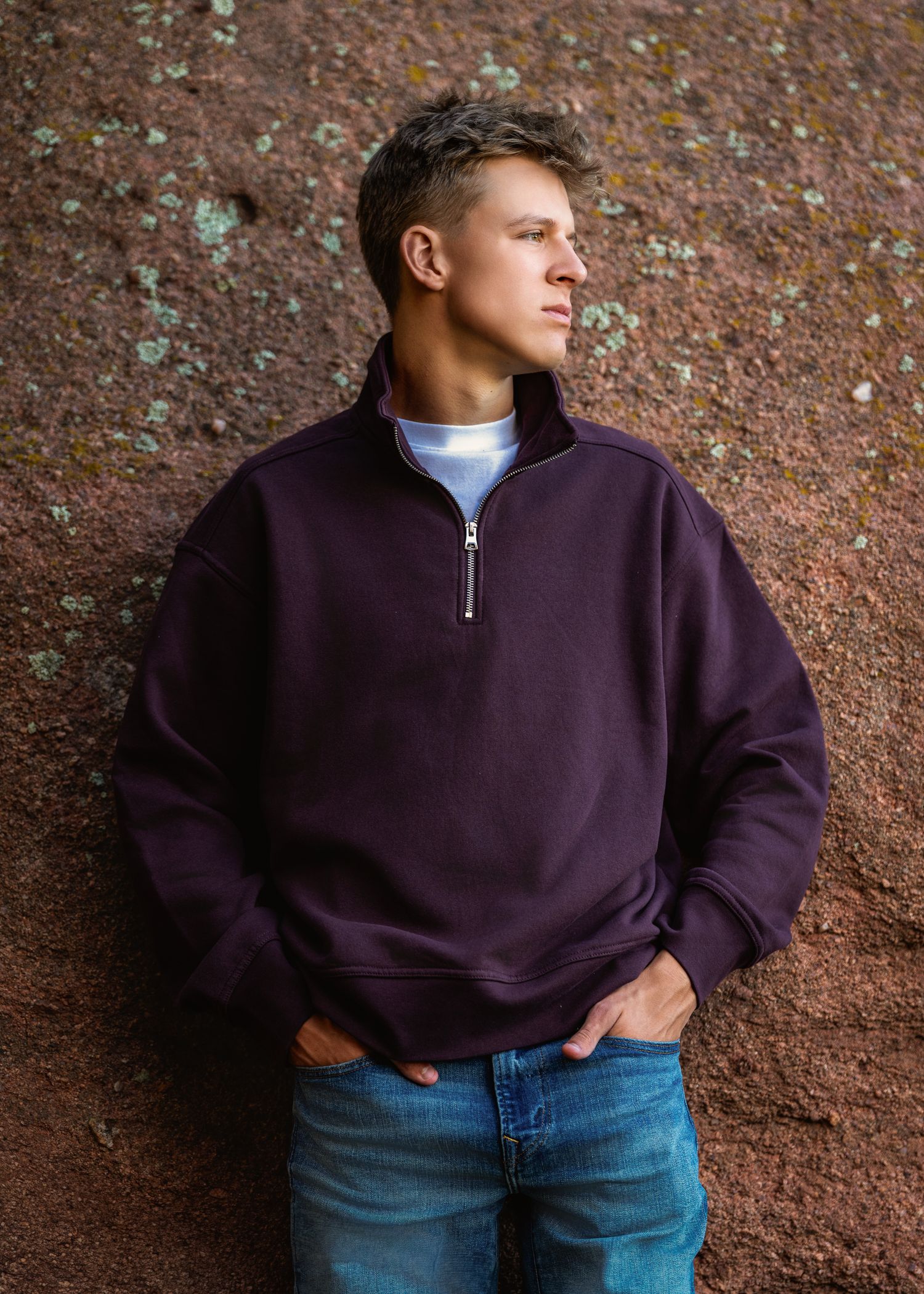 Young man in dark purple quarter-zip sweatshirt and jeans leaning against a textured red rock wall outdoors.