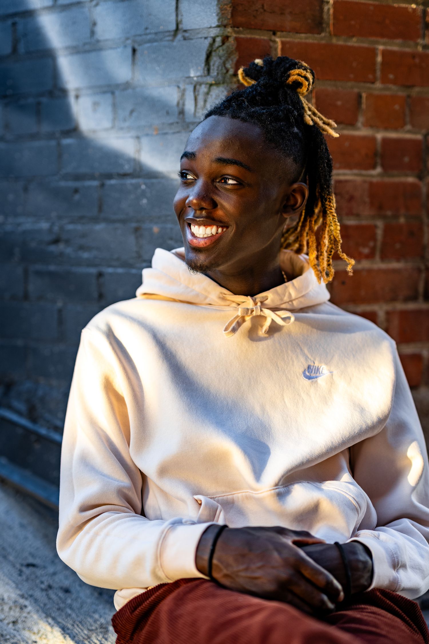 Smiling young Black man with dreadlocks wearing a cream hoodie, sitting against a brick wall in warm sunlight.
