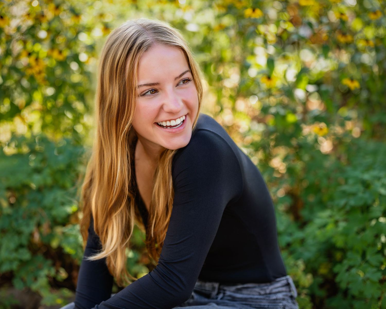 Smiling young blonde woman in black top posing outdoors among lush green foliage in natural light.