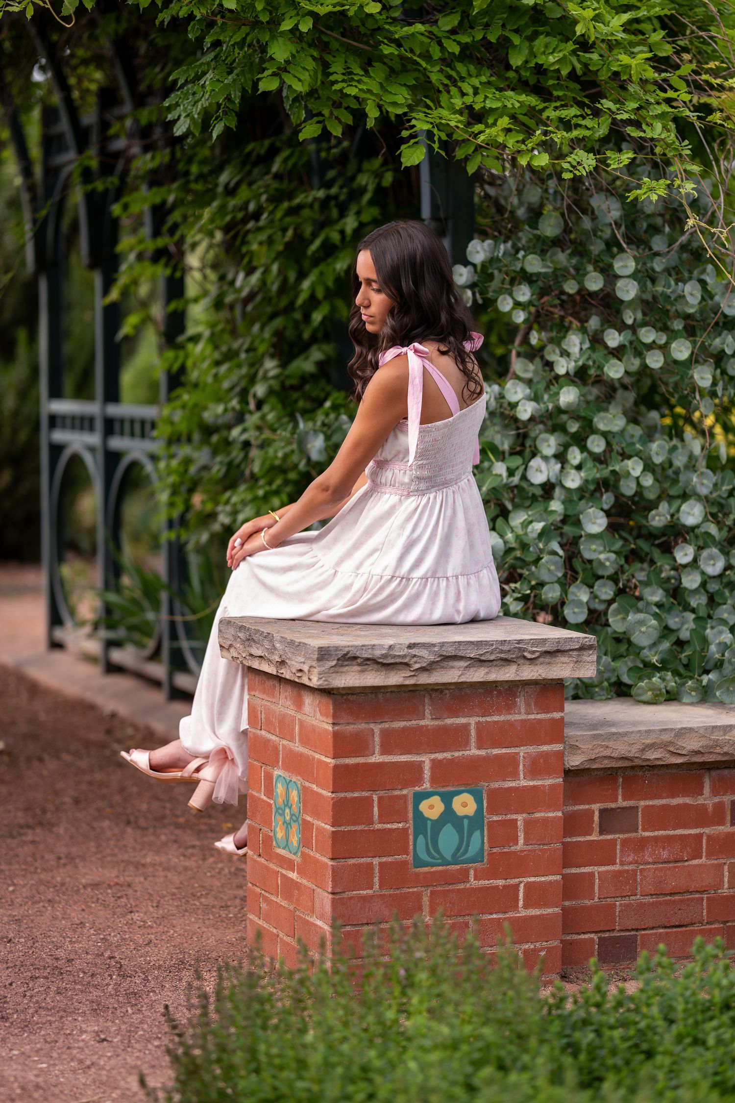 Young woman in white dress sitting on brick pillar in lush garden setting.