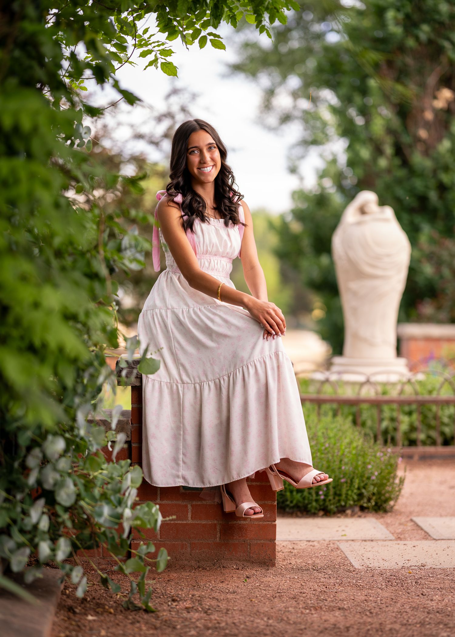 Young woman in white maxi dress sitting in a lush garden with stone sculpture backdrop.
