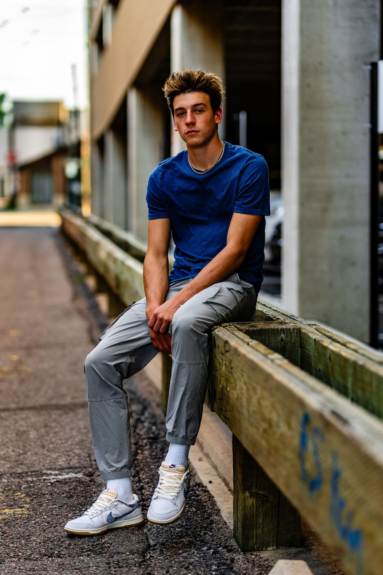 Young man in blue t-shirt and gray pants sitting on wooden ledge in urban alley wearing white sneakers.