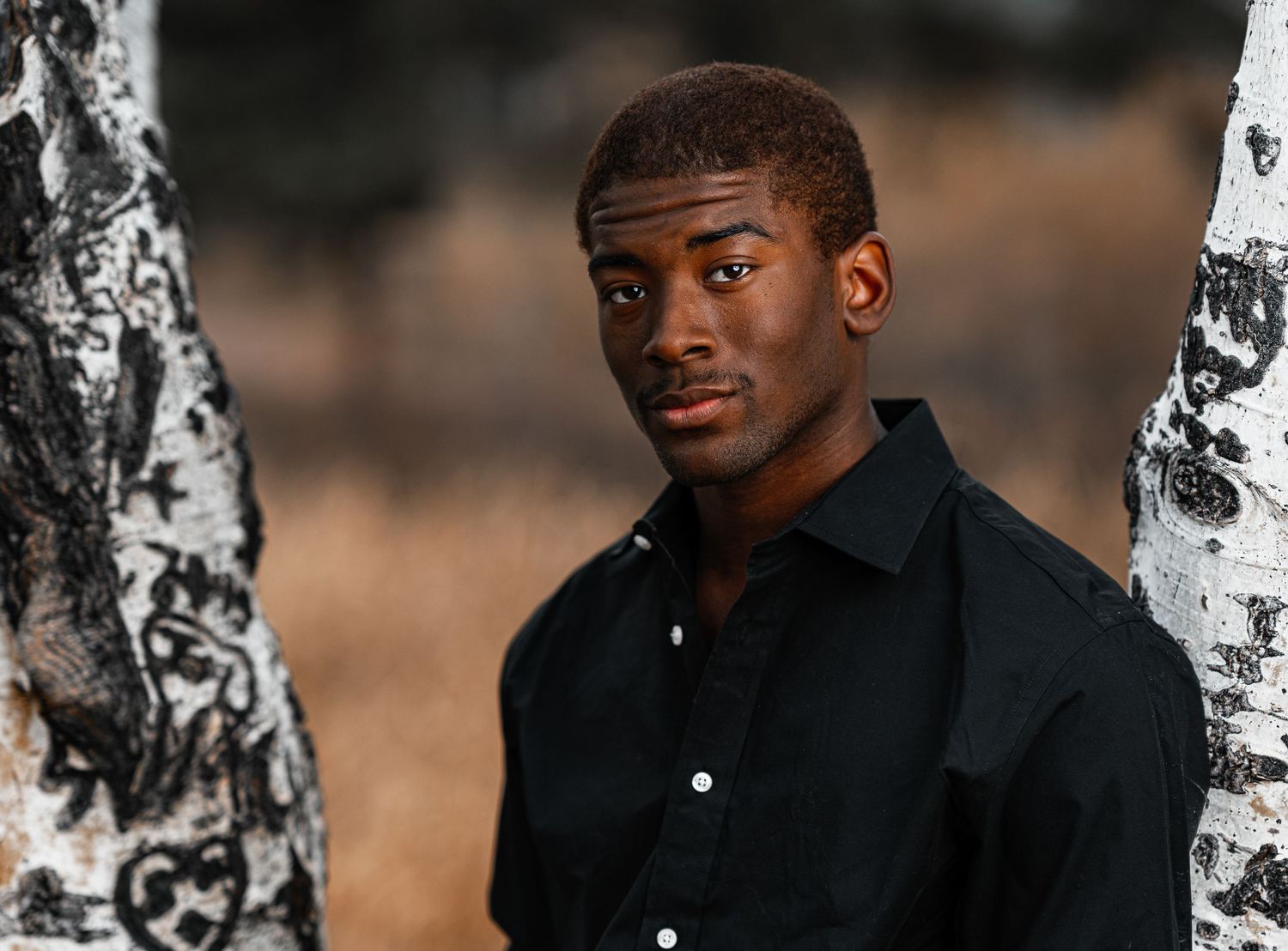 Young Black man in black shirt poses between birch trees in an outdoor portrait photography setting.