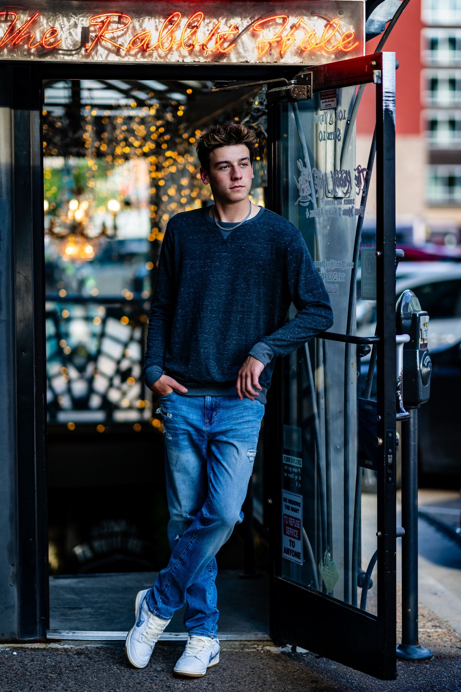 Young man in gray sweater and jeans posing in urban doorway with neon lights and city street background.