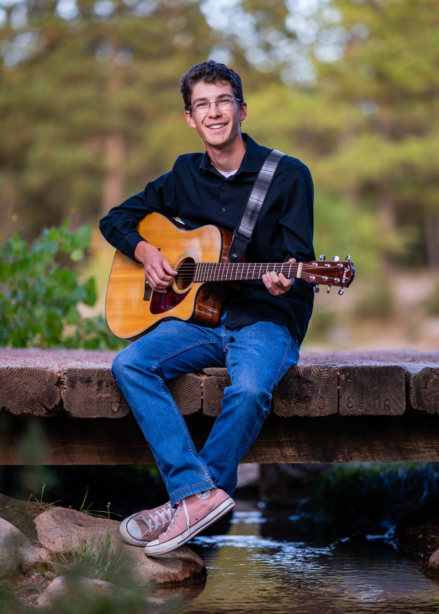 Smiling teenage boy playing acoustic guitar while sitting on a stone bridge outdoors in a scenic park setting.