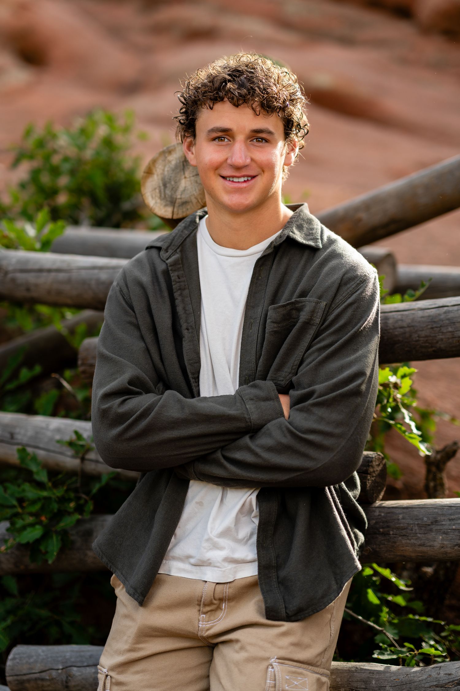 Smiling young man in gray jacket standing arms crossed by rustic log fence outdoors.