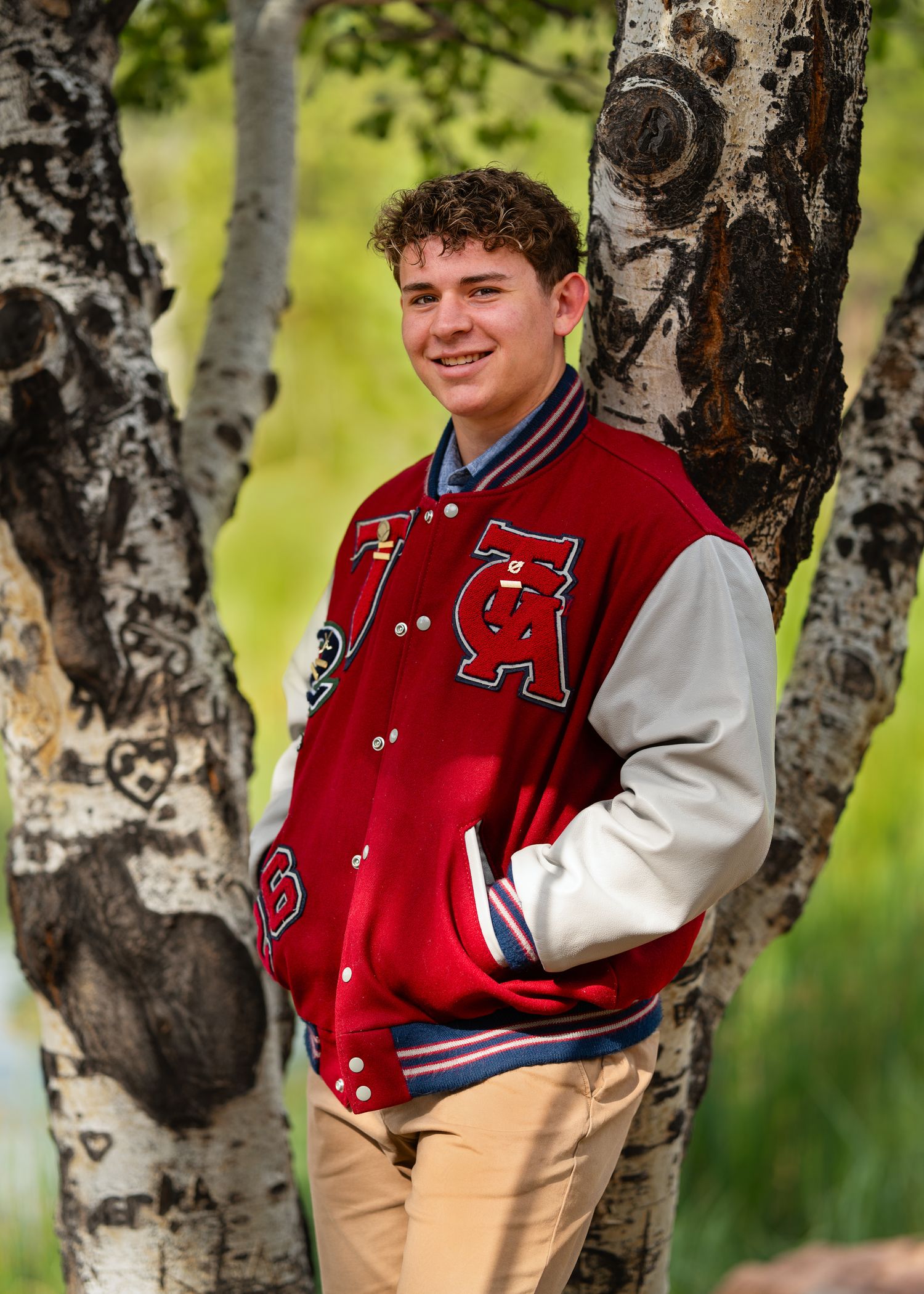 Smiling teen in red varsity letterman jacket leans against birch trees outdoors for senior portrait photo.