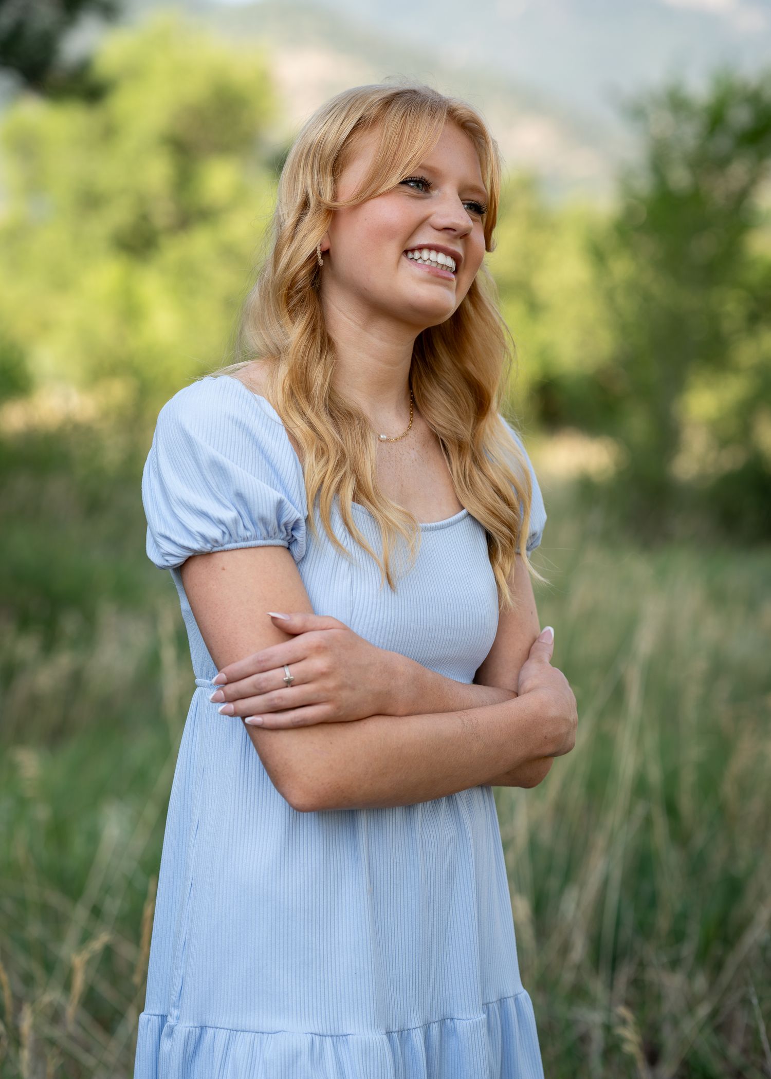 Young blonde woman in light blue dress smiling outdoors in green field with mountains in background.