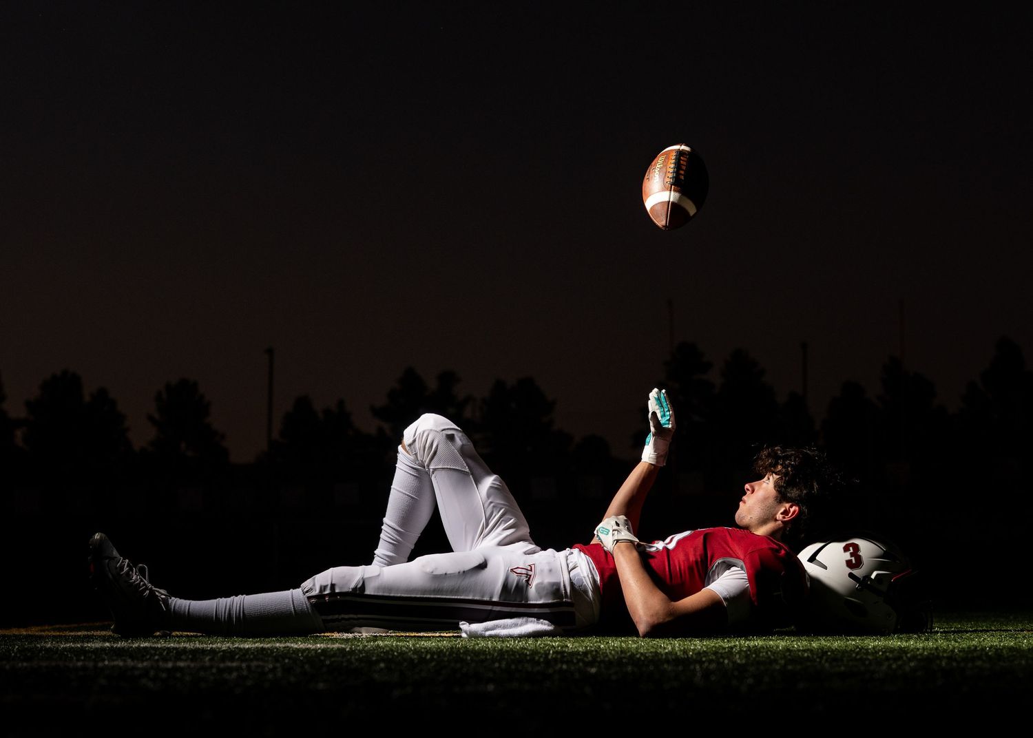 Football player lying on grass at night, reaching up to catch a football under dramatic stadium lighting.