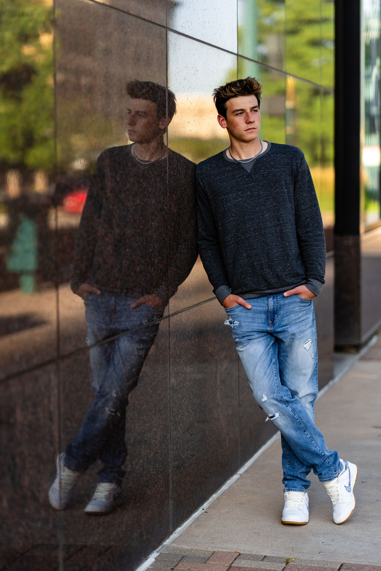 Young man in jeans and dark sweater leaning against reflective black wall, hands in pockets, white sneakers.
