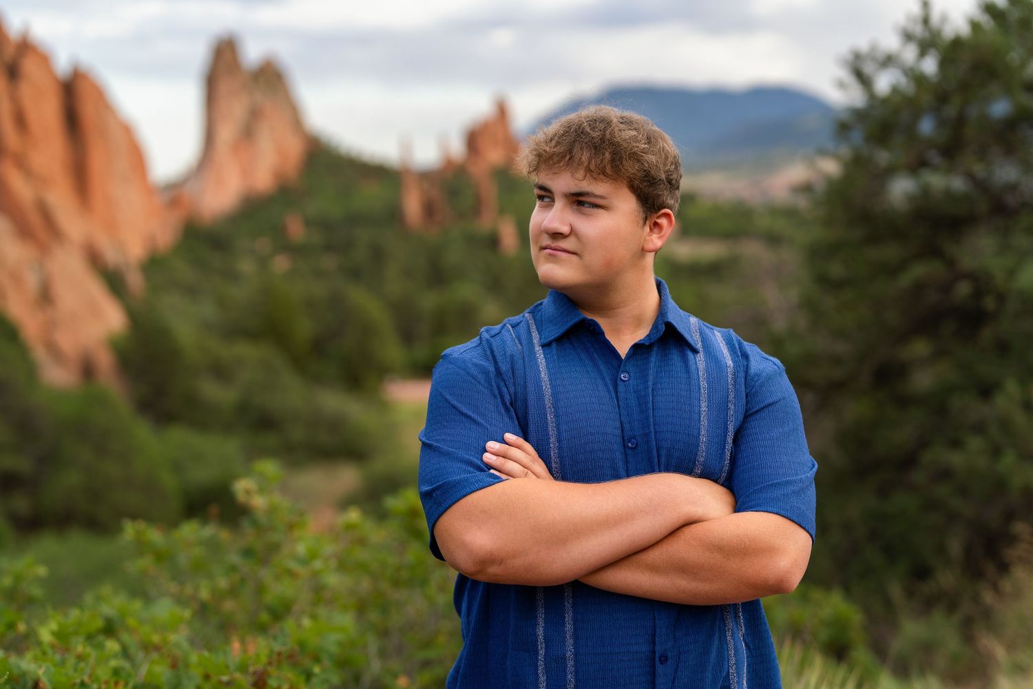 Young man in blue shirt stands with arms crossed at Garden of the Gods, Colorado, with red rock formations behind.