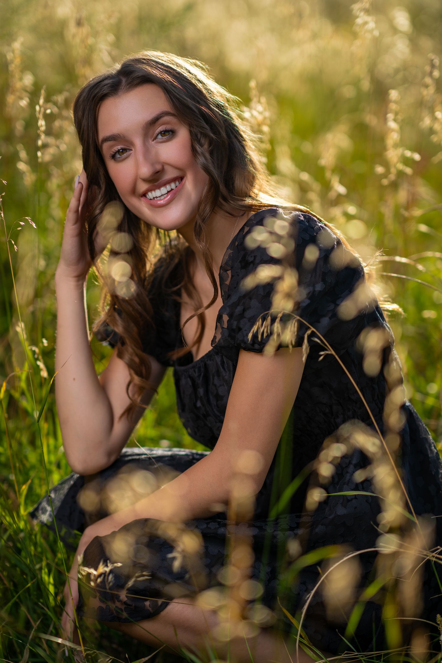 Smiling young woman in black tie-dye dress sitting in tall green grass during golden hour.