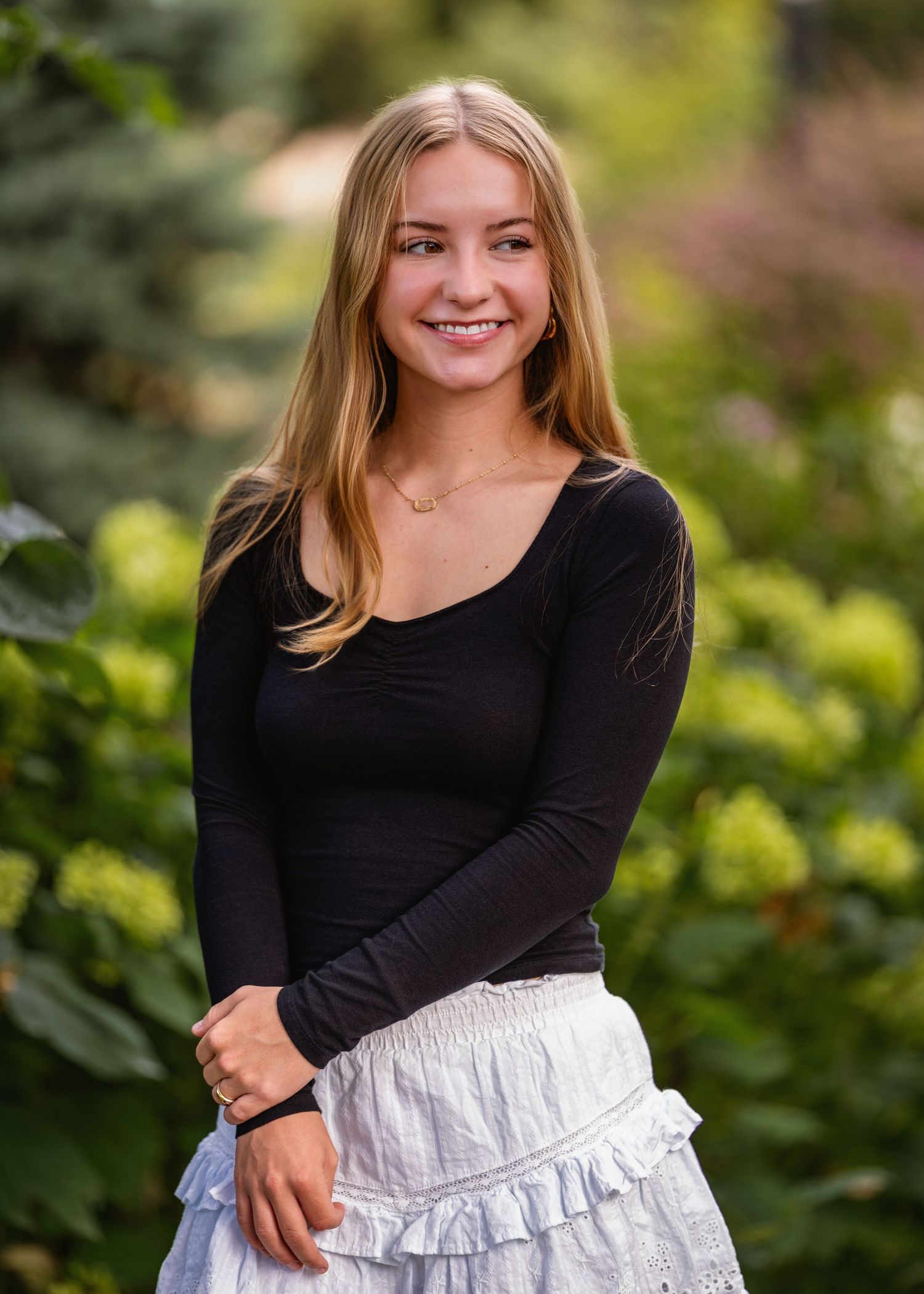 Smiling young woman with long blonde hair wearing a black top and white ruffled skirt, posing outdoors among lush green foliage.
