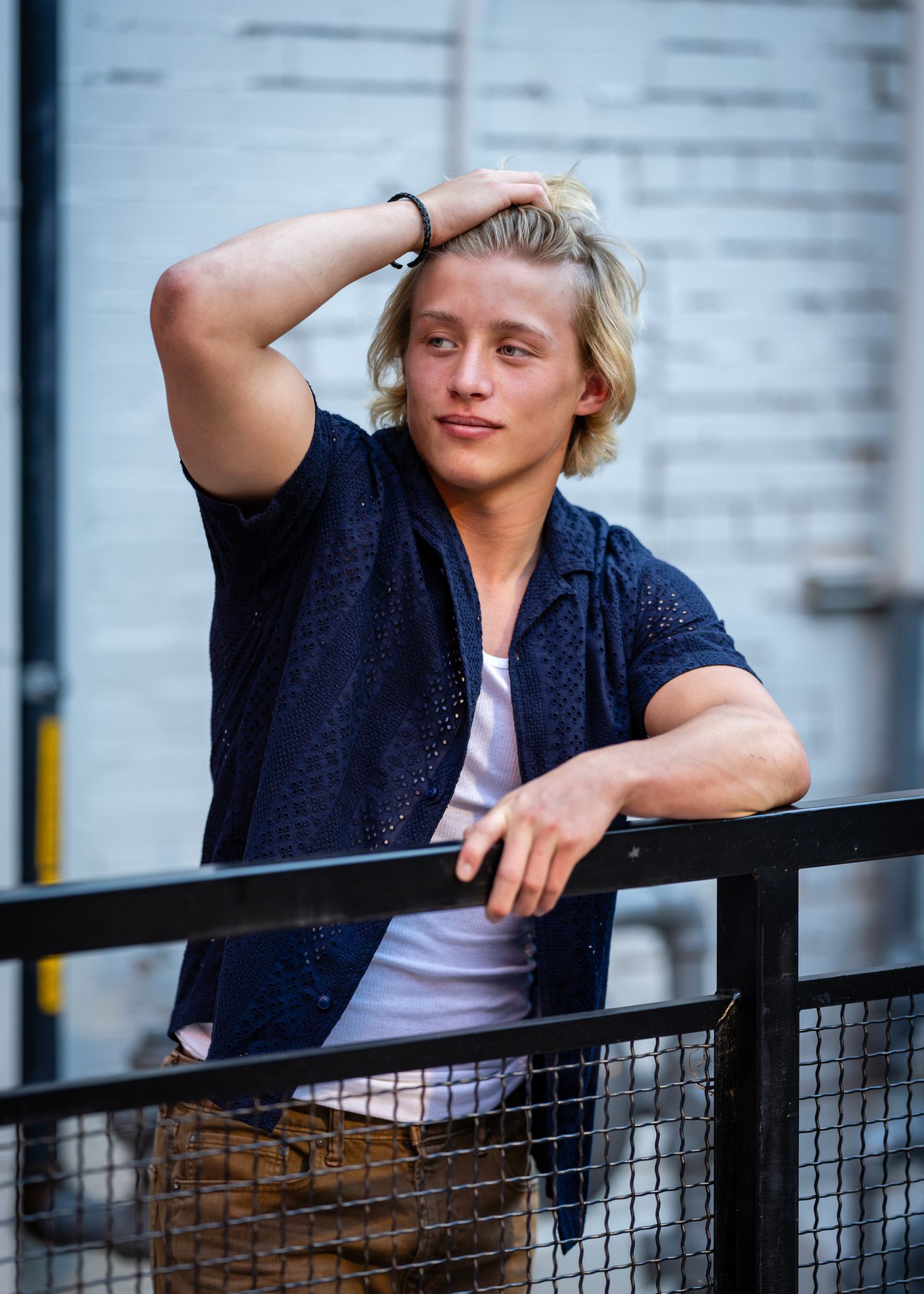 Blonde young man in navy shirt leaning on black metal railing with hand in hair.