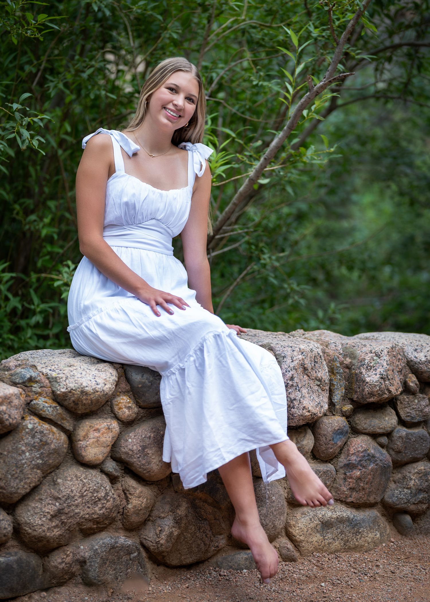 Smiling young woman in white sundress sitting barefoot on stone wall surrounded by lush green trees.