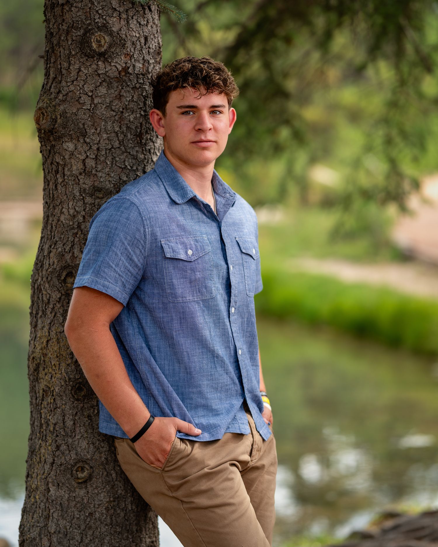 Young man in blue shirt leaning against tree near river for outdoor senior portrait photo.