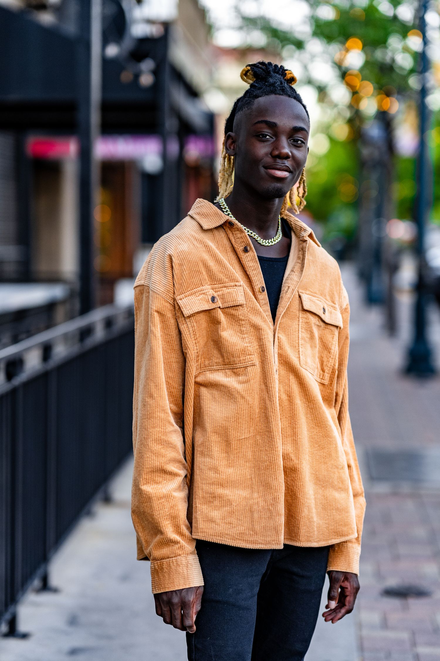 Young Black man in tan corduroy jacket and black jeans posing on urban sidewalk with bokeh city background.