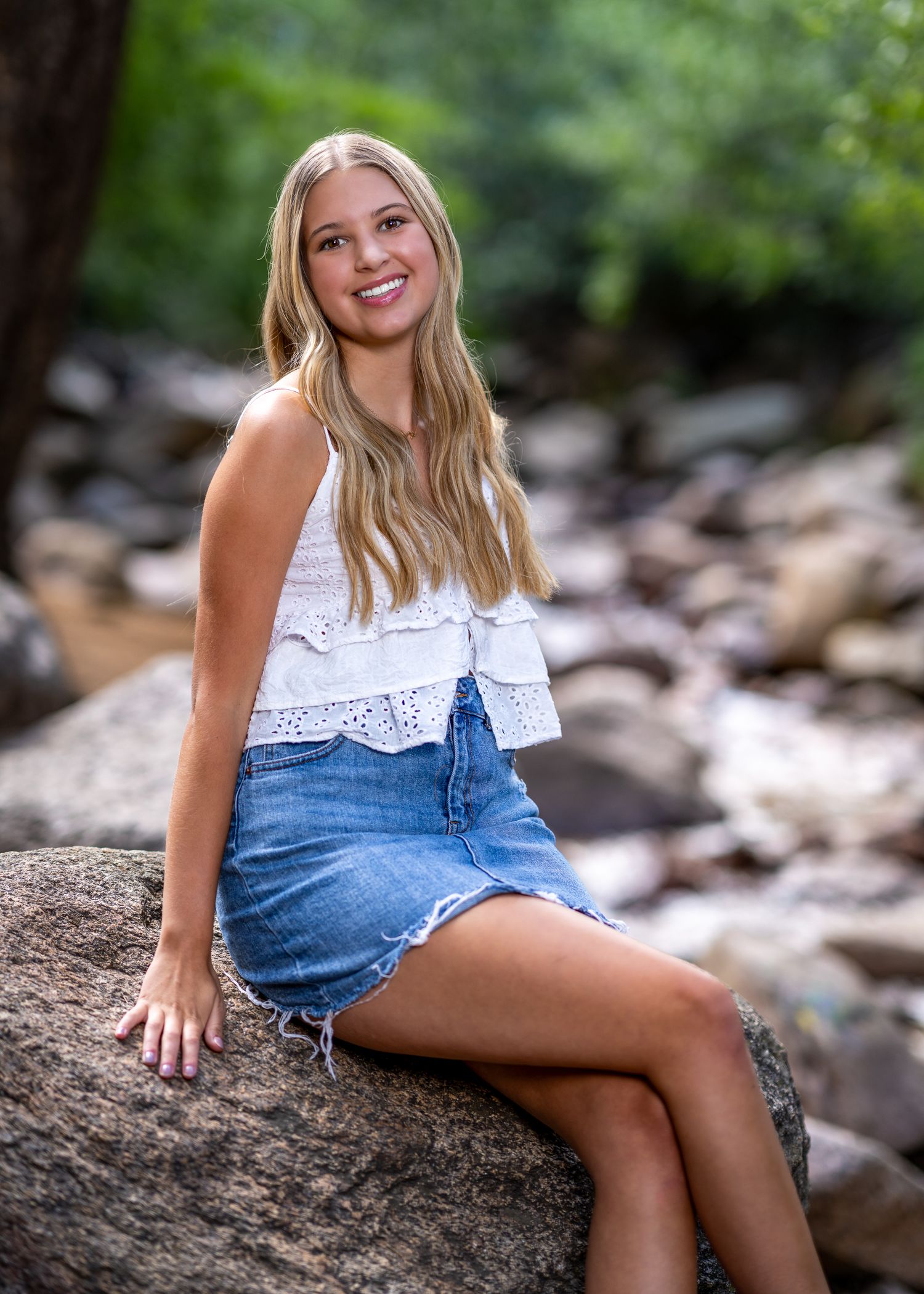 Smiling teenage girl in white crop top and denim skirt sitting on rocks near a stream outdoors.