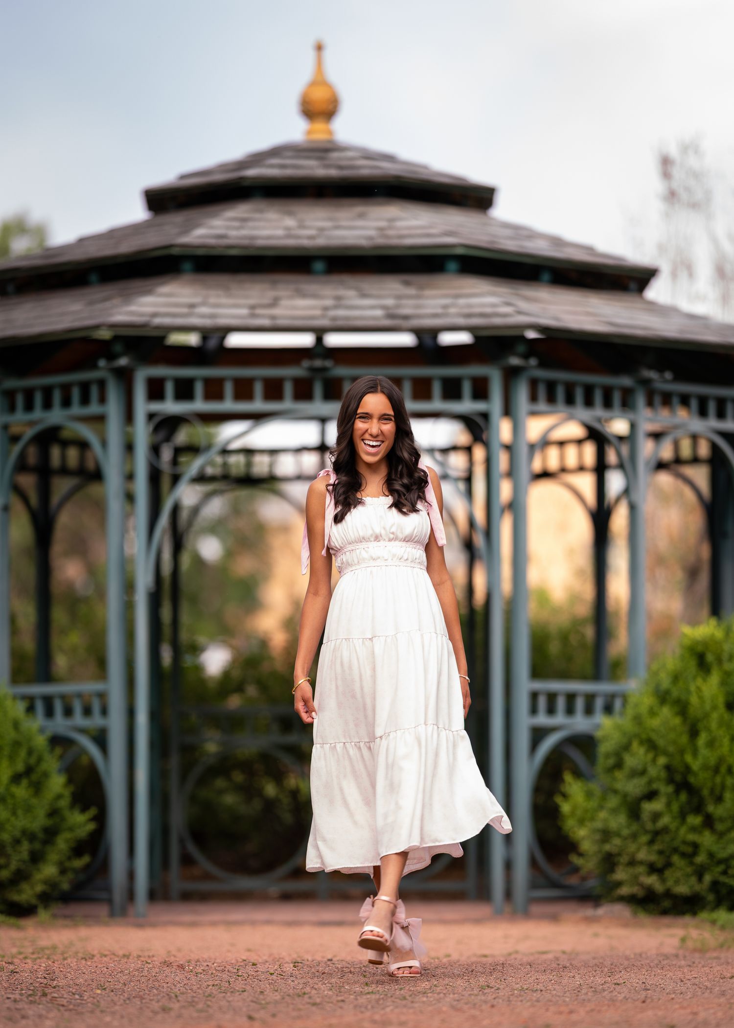 A smiling woman in a white maxi dress poses in front of a decorative teal gazebo in a lush garden setting.