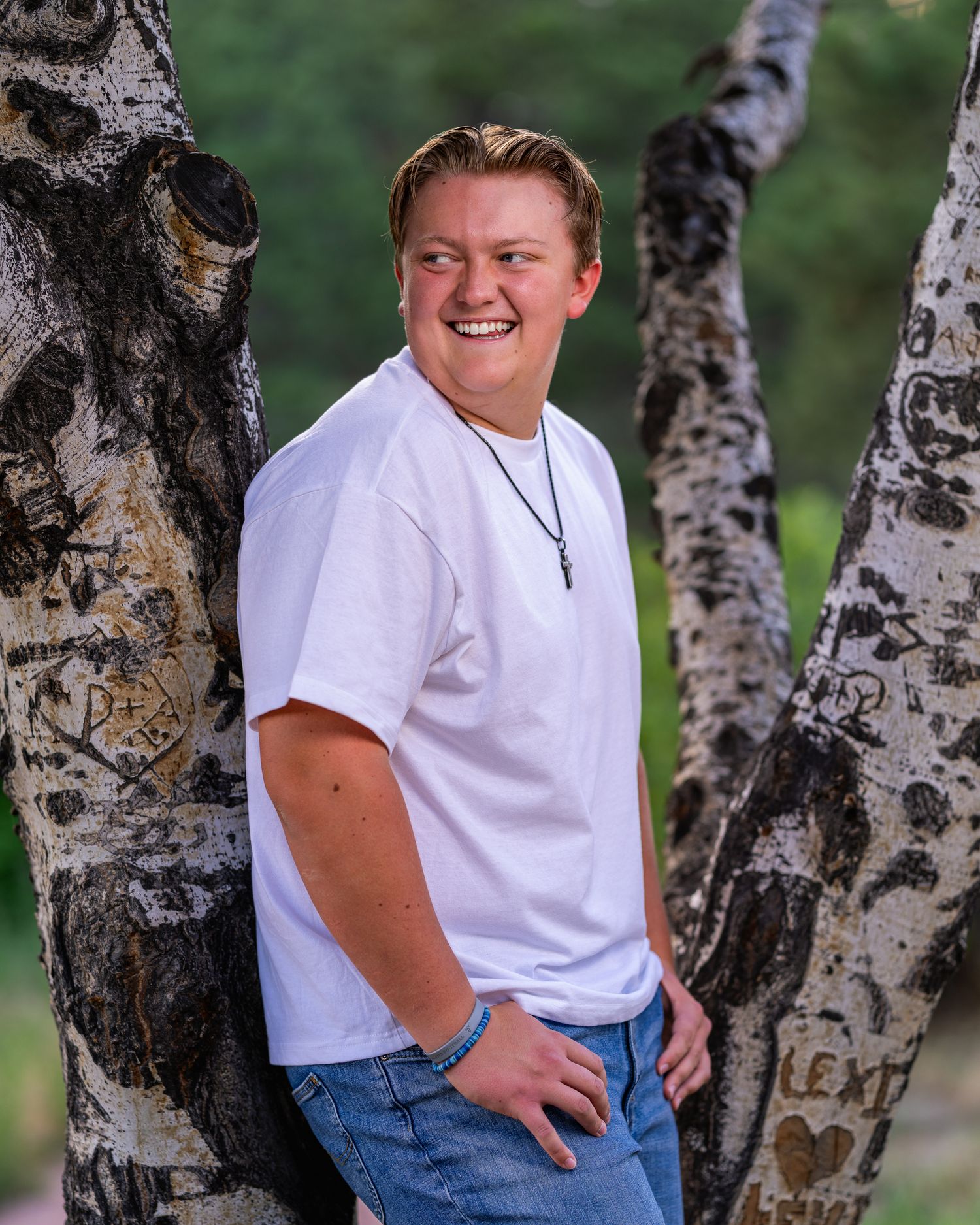 Smiling young man in white t-shirt leaning against birch trees with carvings, wearing a cross necklace outdoors.