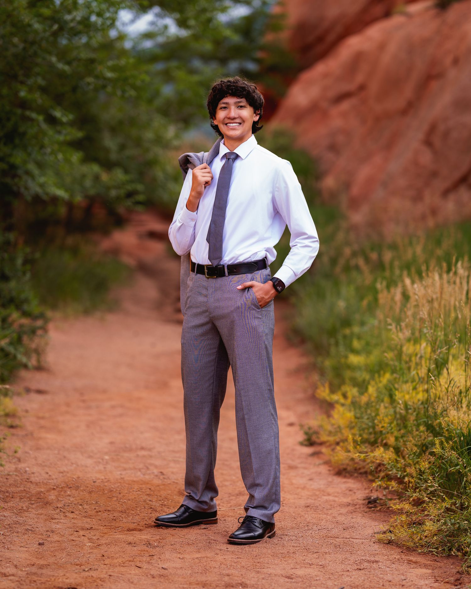 Young man in white dress shirt, tie, and gray slacks poses on a red dirt path surrounded by lush greenery.