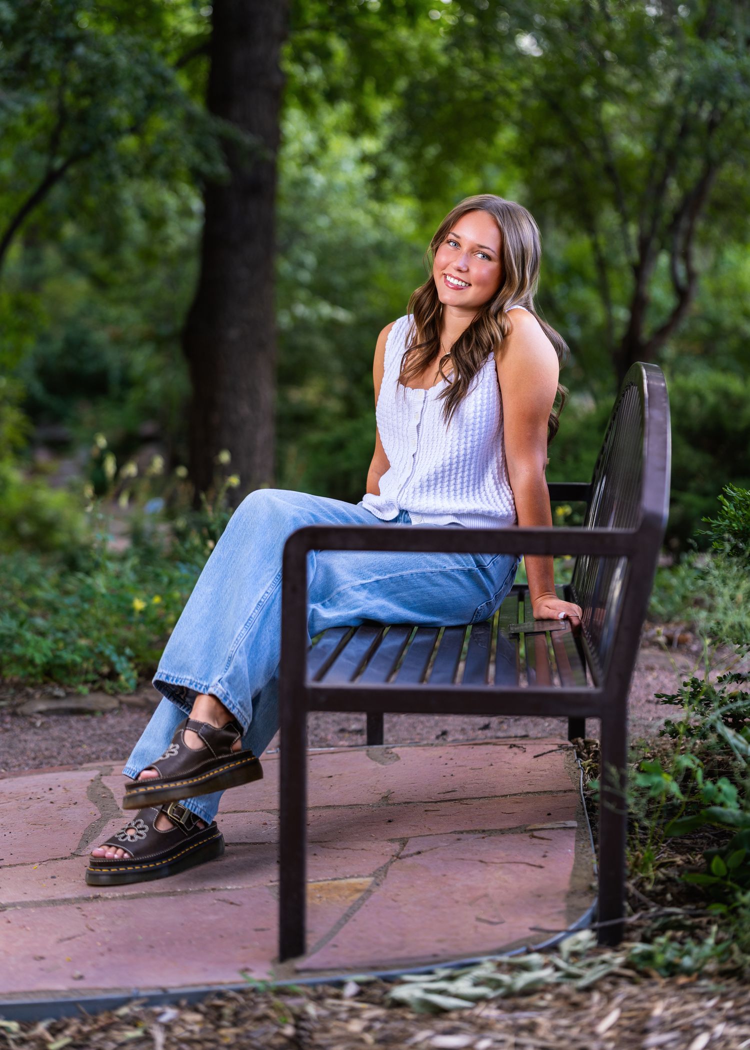 Young woman smiling on a park bench wearing jeans and white top, surrounded by lush greenery.