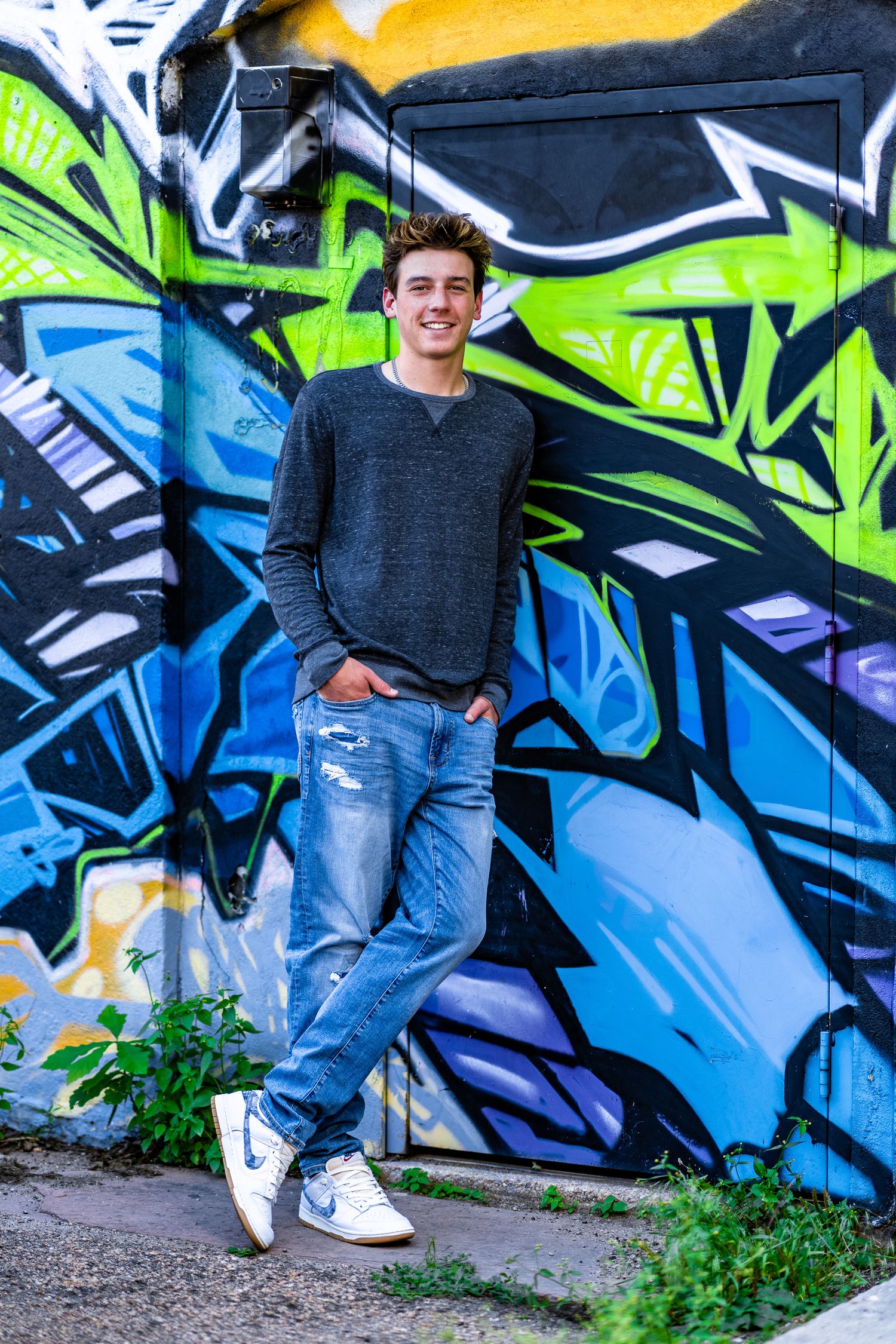 Smiling young man in gray sweater and ripped jeans leaning against a colorful graffiti wall outdoors.