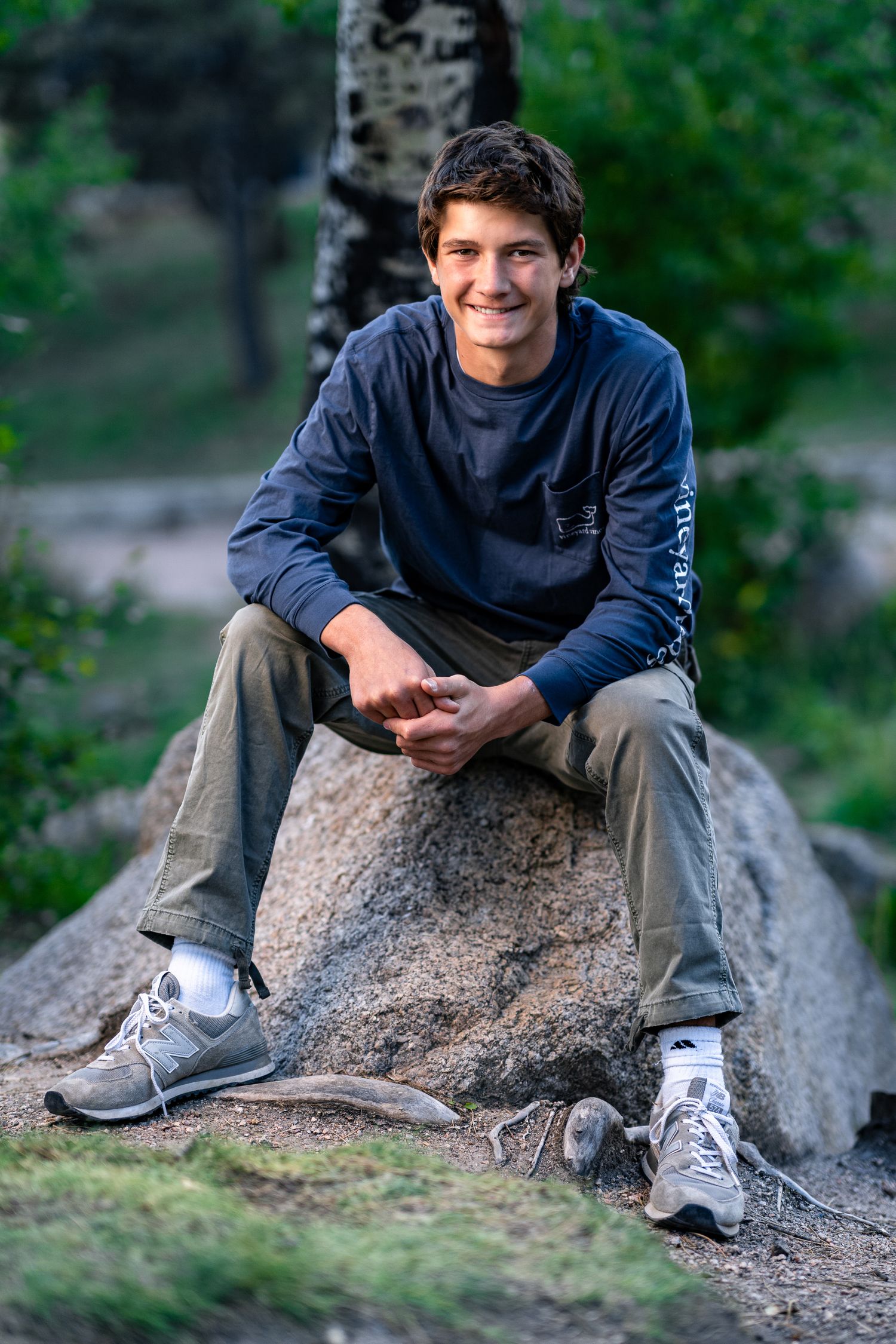 Smiling teenage boy sitting on a boulder outdoors in a forest, wearing a navy long-sleeve shirt and gray pants.