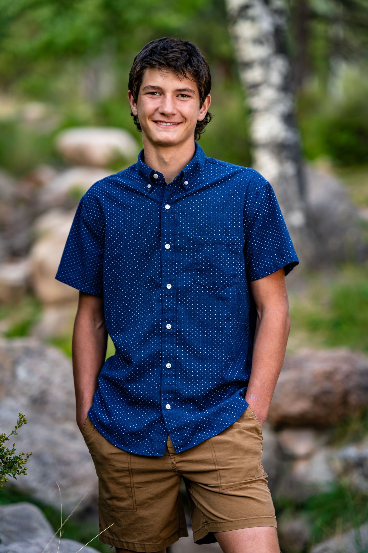 Smiling teenage boy in blue patterned shirt and khaki shorts posing outdoors among rocks and trees.