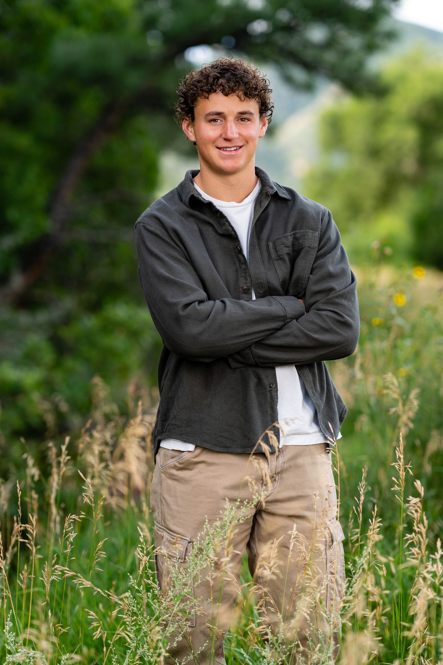 Young man with curly hair stands smiling in a field, arms crossed, wearing a dark jacket and khaki pants.