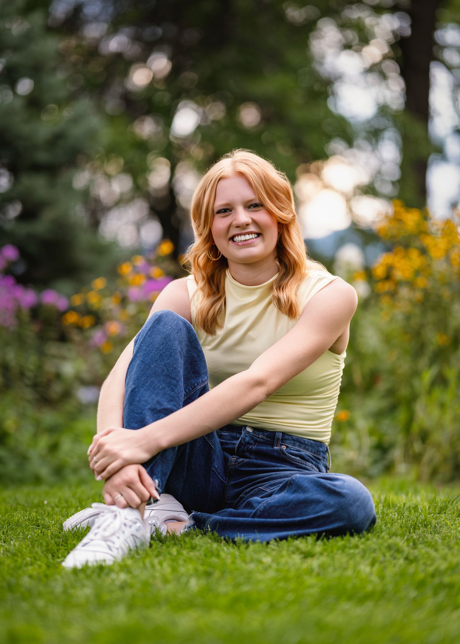 Smiling young woman with red hair sitting on green grass in a garden wearing a yellow top and jeans.