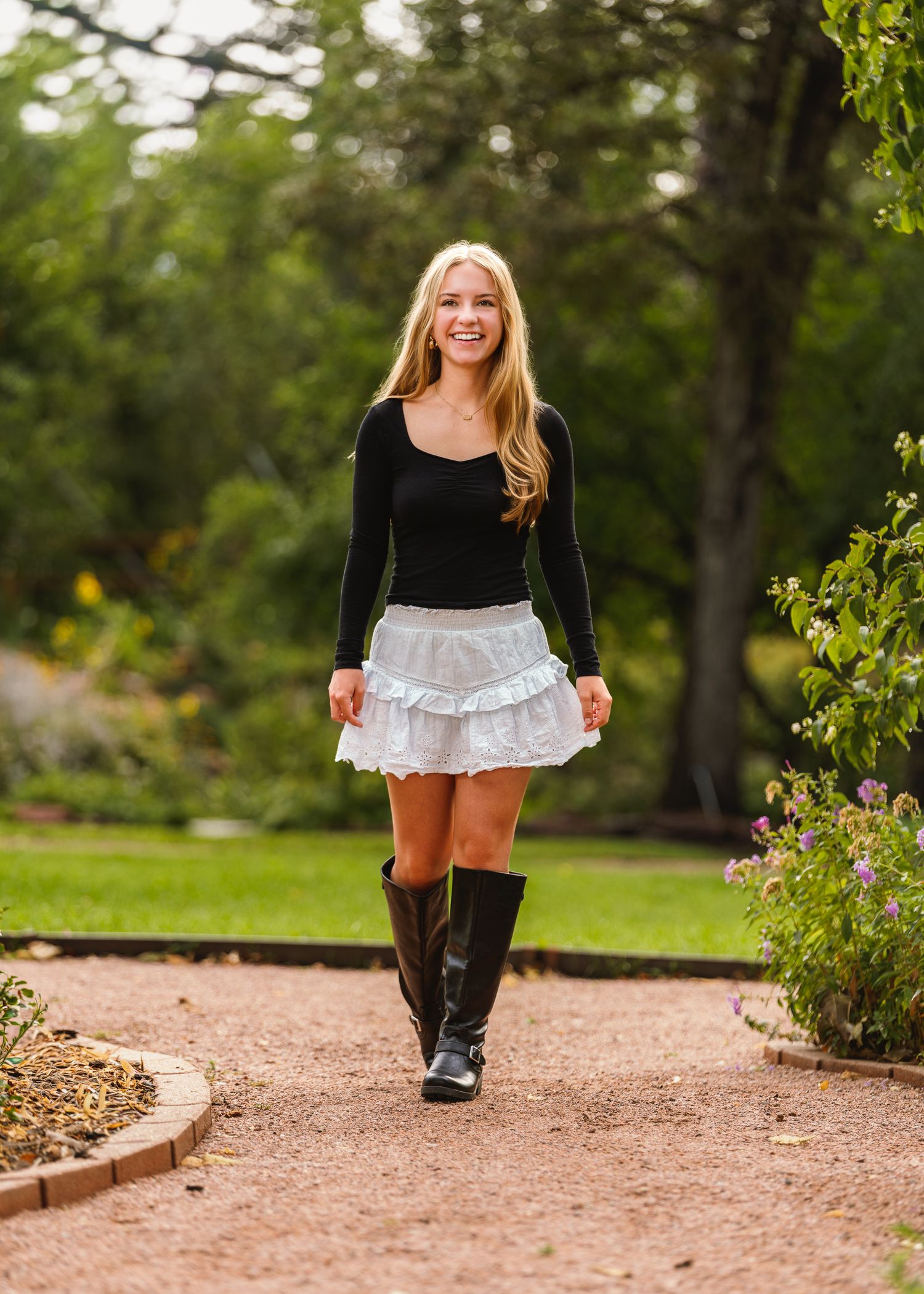 Smiling blonde woman in black top, white ruffled mini skirt, and knee-high boots walking on a garden path.