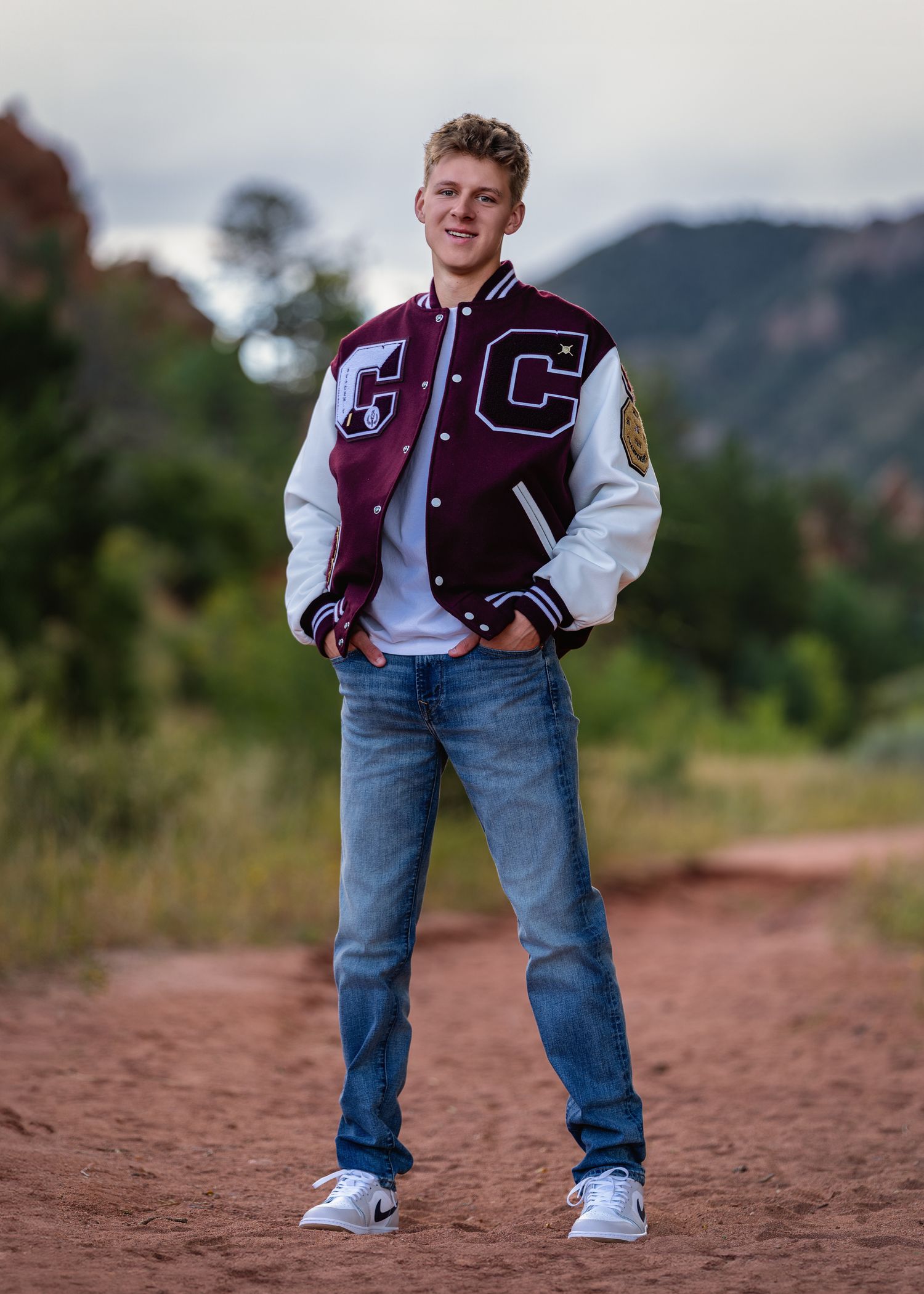 Teen boy in varsity jacket and jeans poses on red dirt path with mountain backdrop for senior photos.