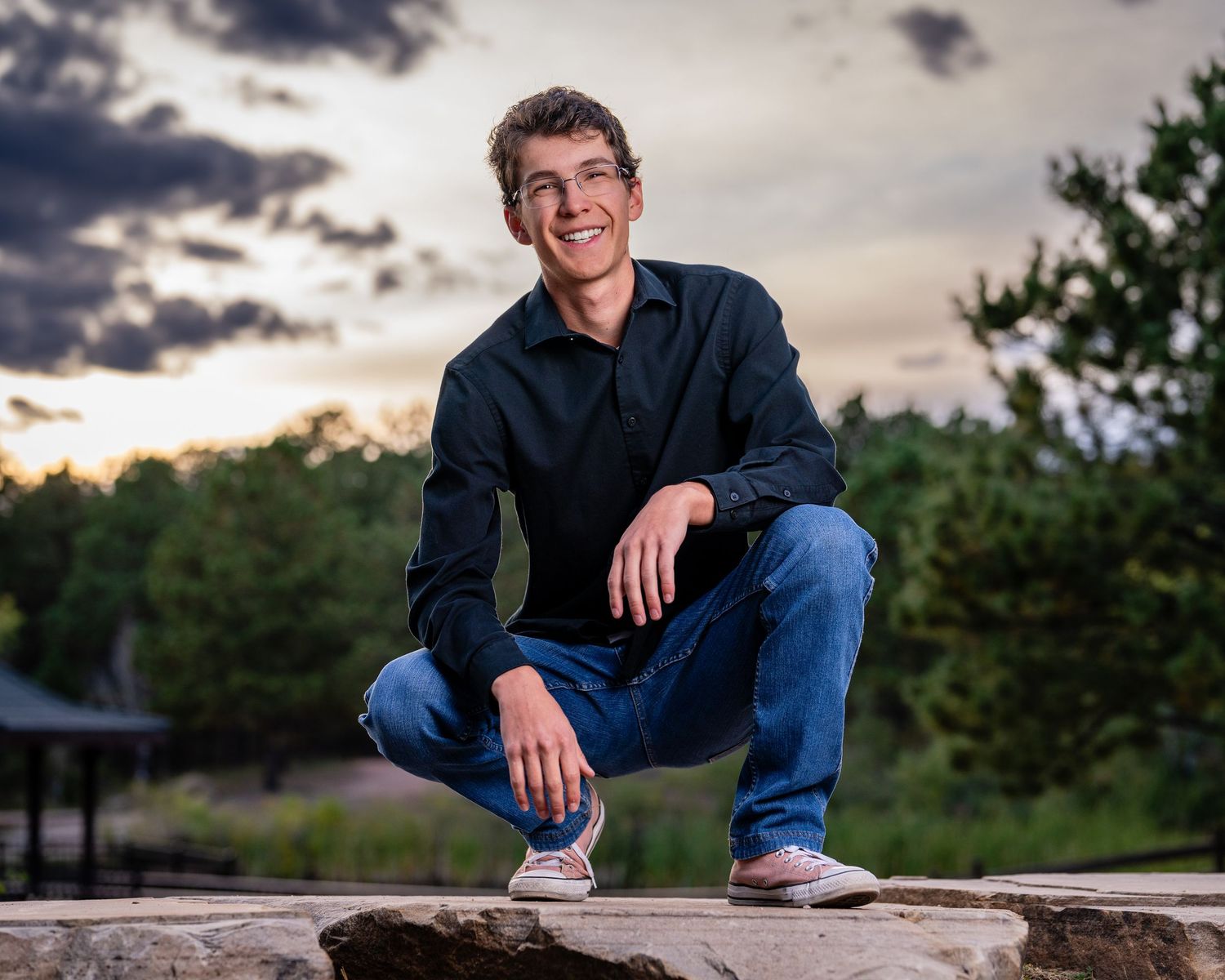Young man in black shirt and jeans crouching on stone wall, smiling outdoors at sunset with trees behind him.