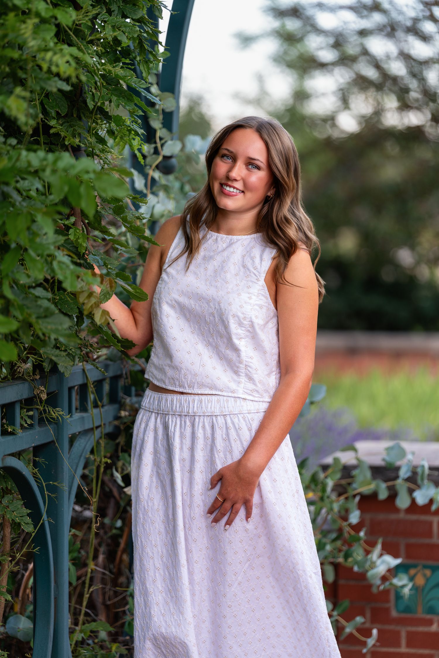 Smiling young woman in white dress posing by ivy-covered garden trellis outdoors.