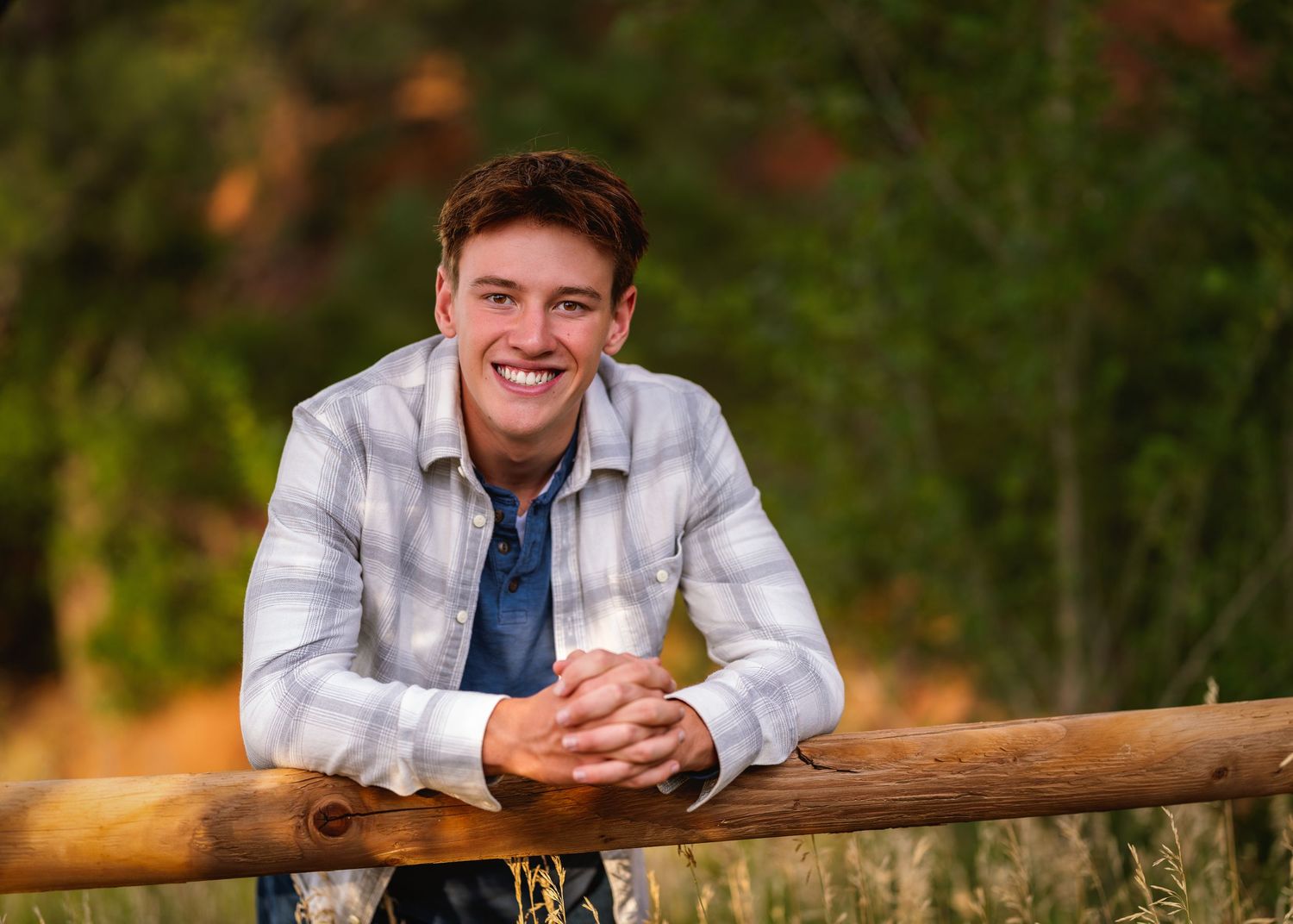 Smiling young man in plaid shirt leaning on rustic wooden fence rail outdoors with colorful autumn foliage background.
