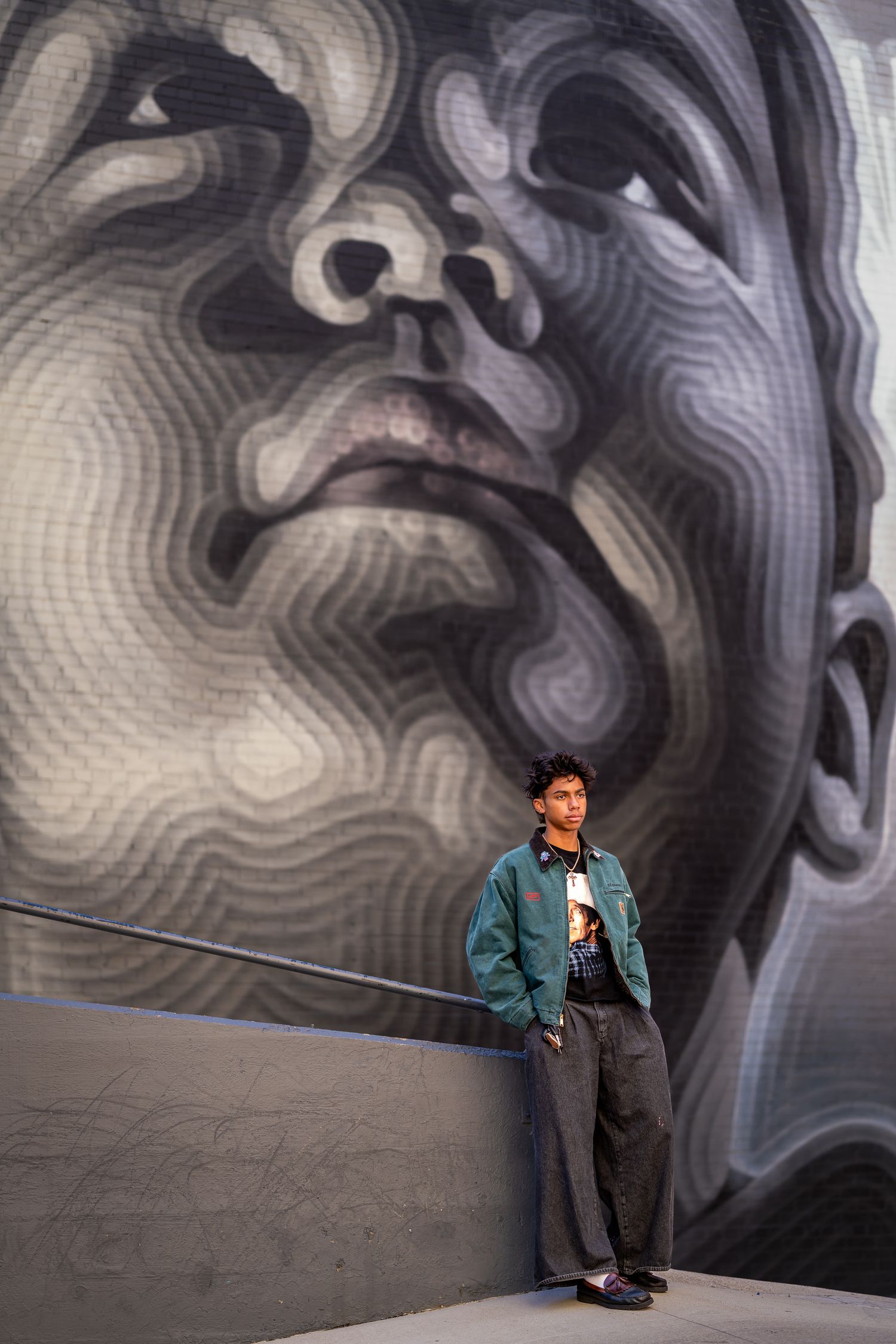 Young man in teal jacket stands before a large black and white mural of a face on a gray wall.