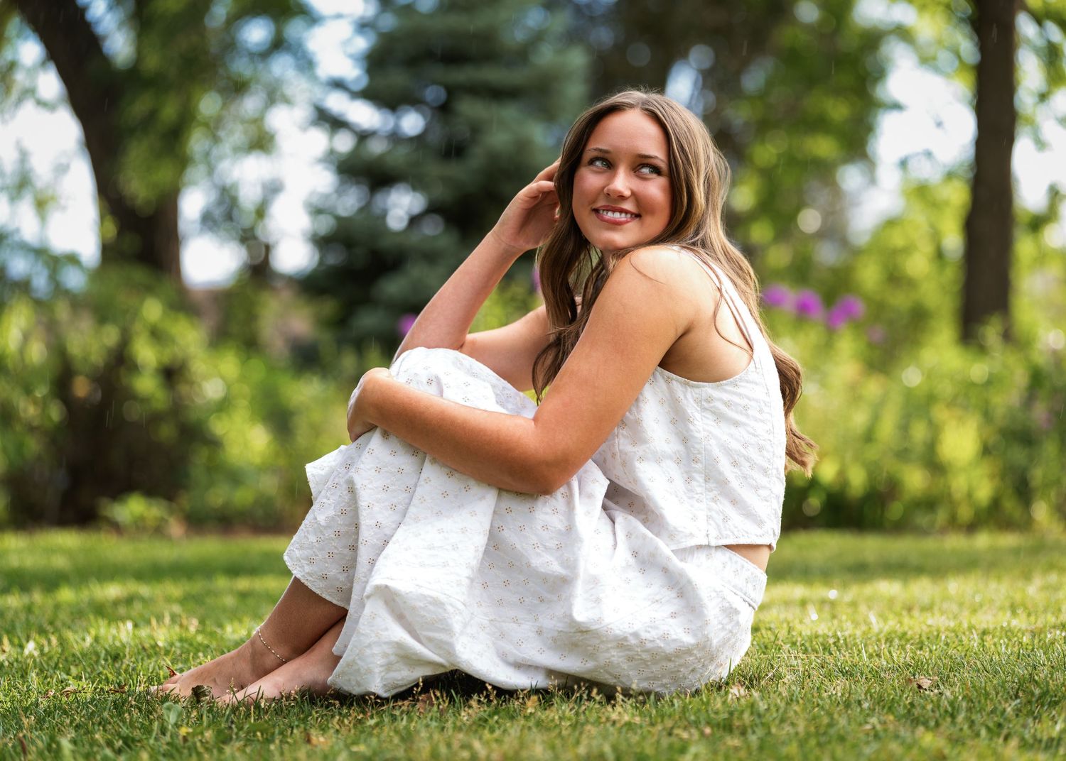 Young woman in white dress sitting on green grass in a sunny garden, smiling and posing for a portrait.