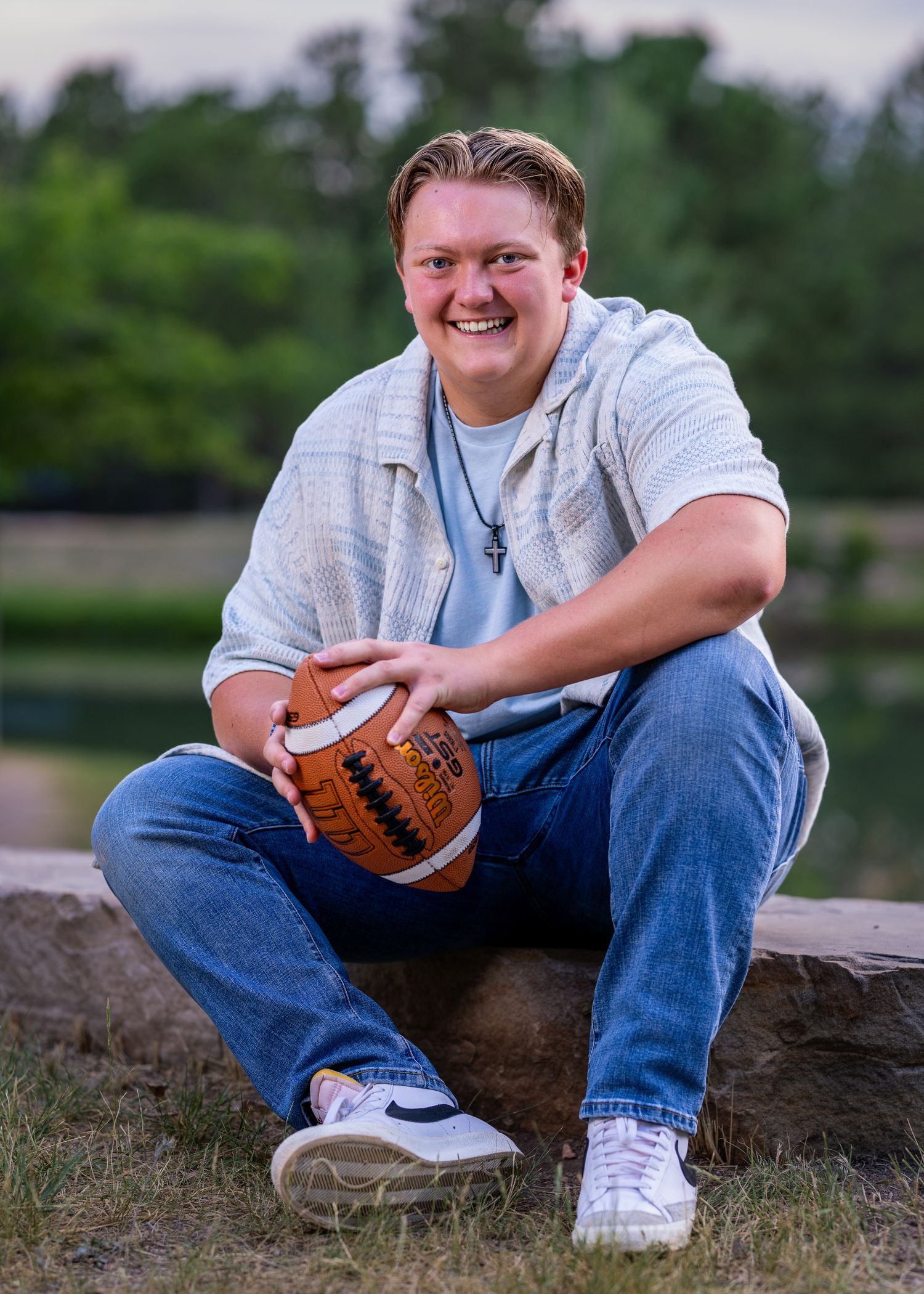Smiling young man sitting on a rock holding a signed football, wearing jeans and a light blue shirt outdoors.