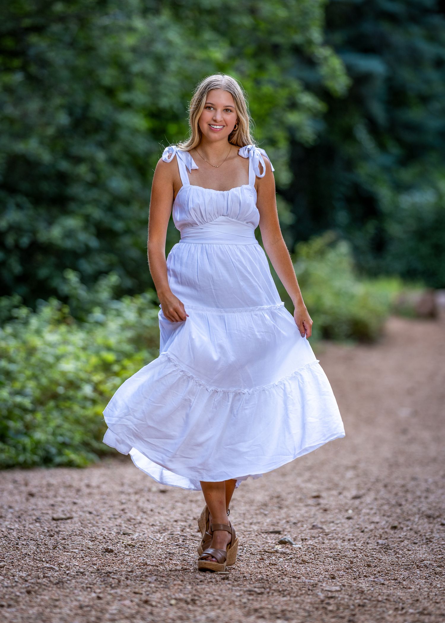 Young blonde woman walking in a white tiered midi dress with tie shoulders on a dirt path surrounded by greenery.