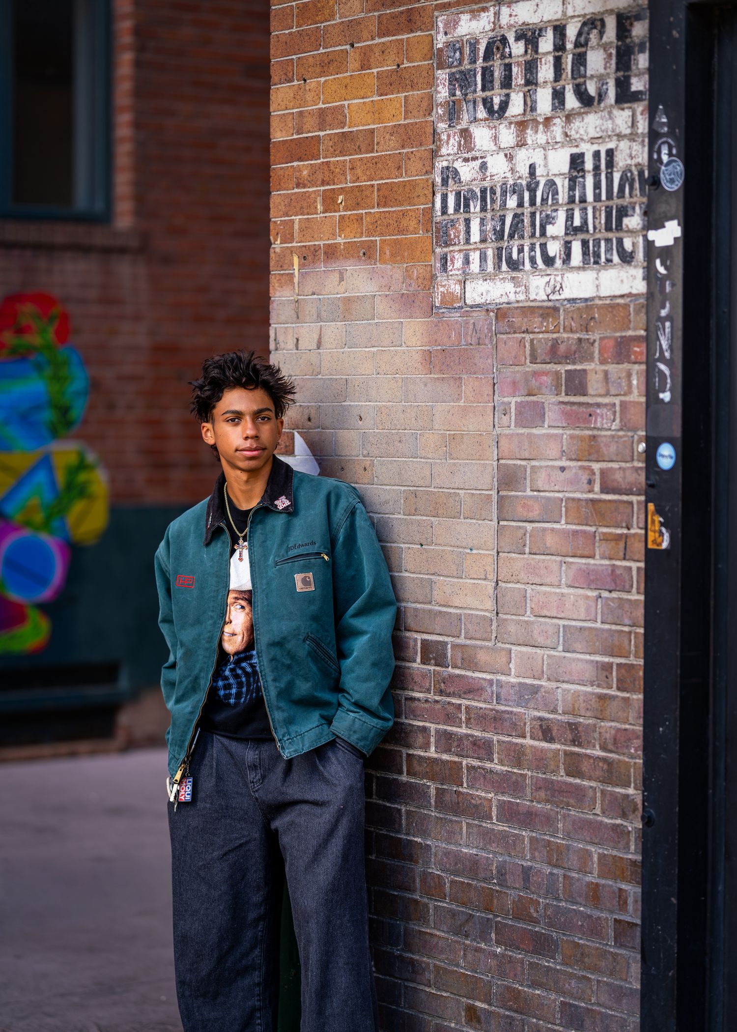 Young man in teal Carhartt jacket leaning against brick wall near a Private Alley sign.