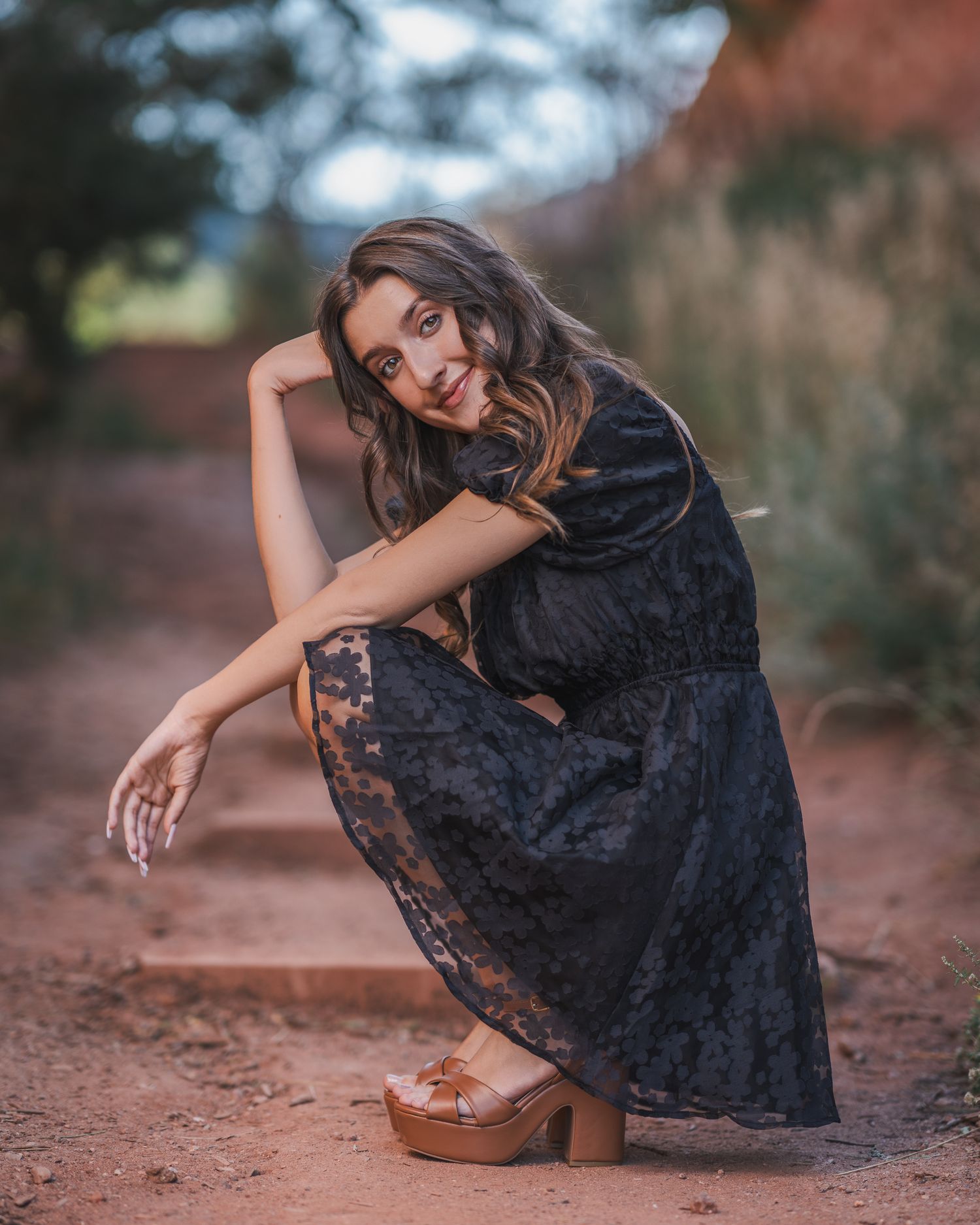 Young woman in black lace dress and wooden platform heels posing on a red dirt path with blurred greenery background.