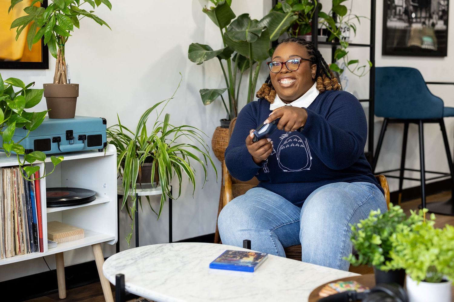 A smiling Black woman with glasses sits in a cozy living room holding a camera, surrounded by plants and a vinyl record player.