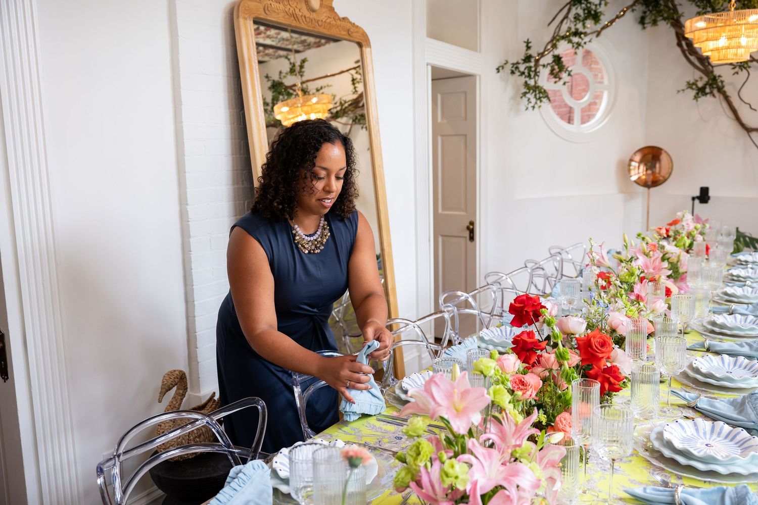 Black woman in navy sleeveless dress setting elegant floral dining table with gold mirror behind.