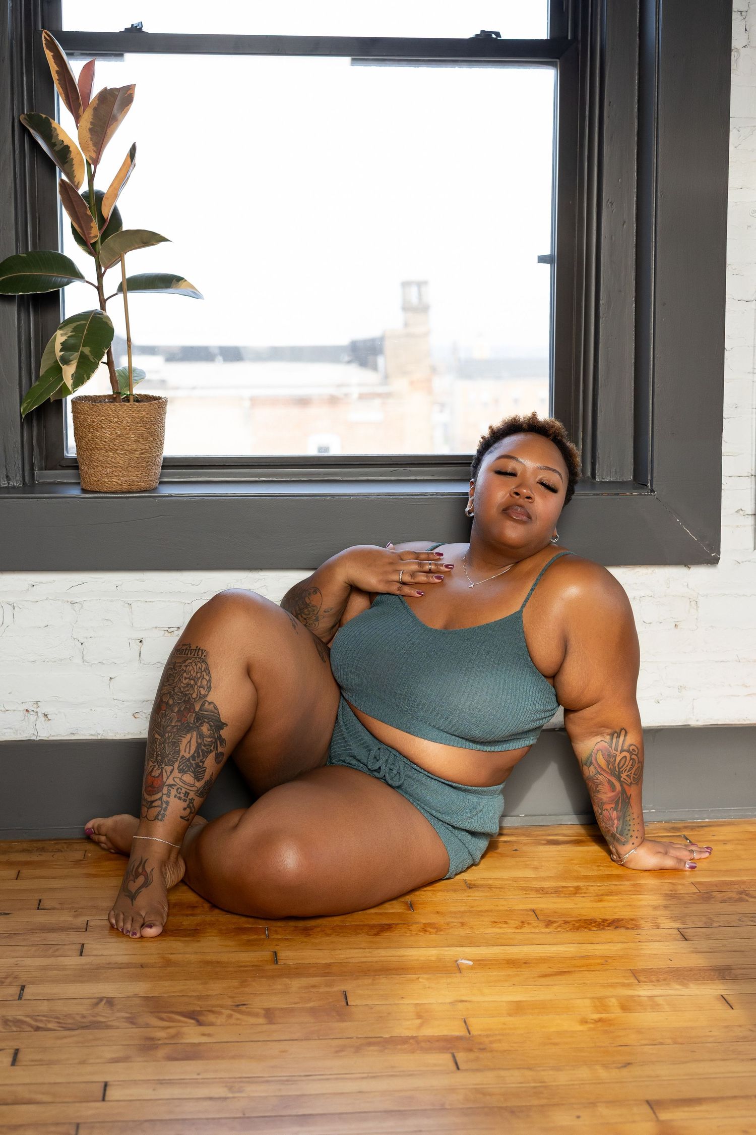 Plus-size woman with tattoos poses confidently in teal crop top and shorts on hardwood floor by window.