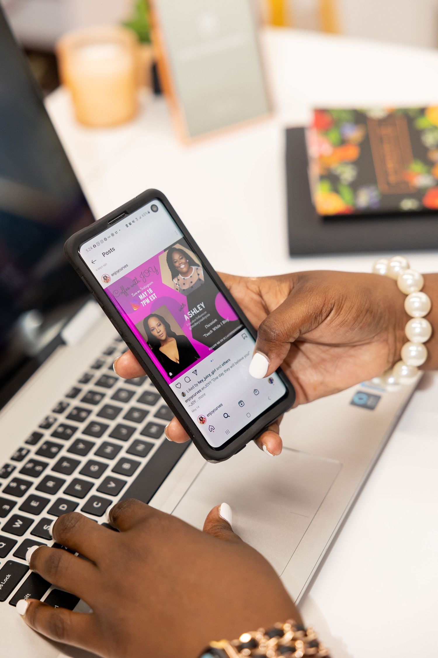 Black woman holding smartphone displaying social media profile while working at laptop with pearl bracelet.